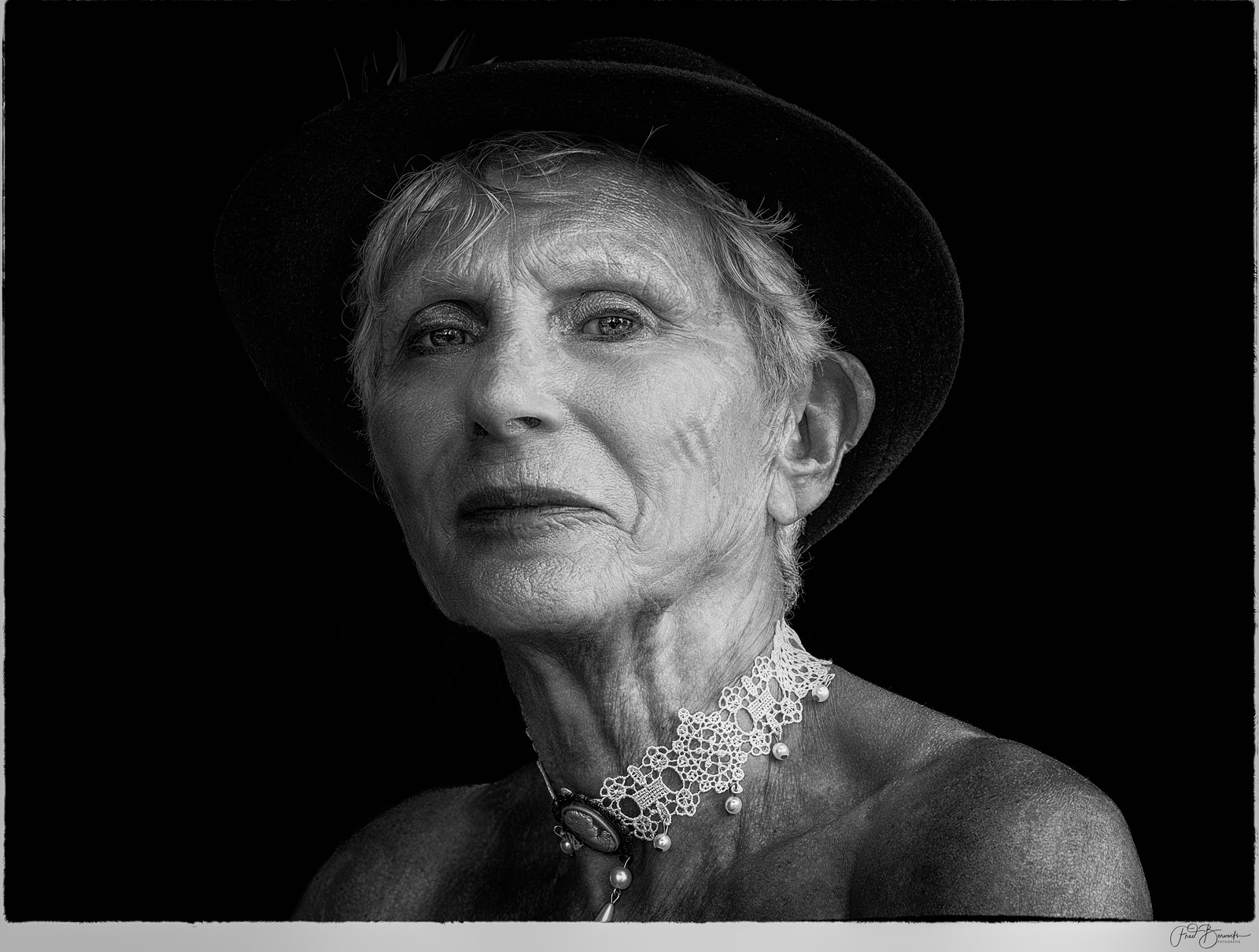 An elderly woman in a hat and lace collar is shown in a close-up portrait against a dark background.