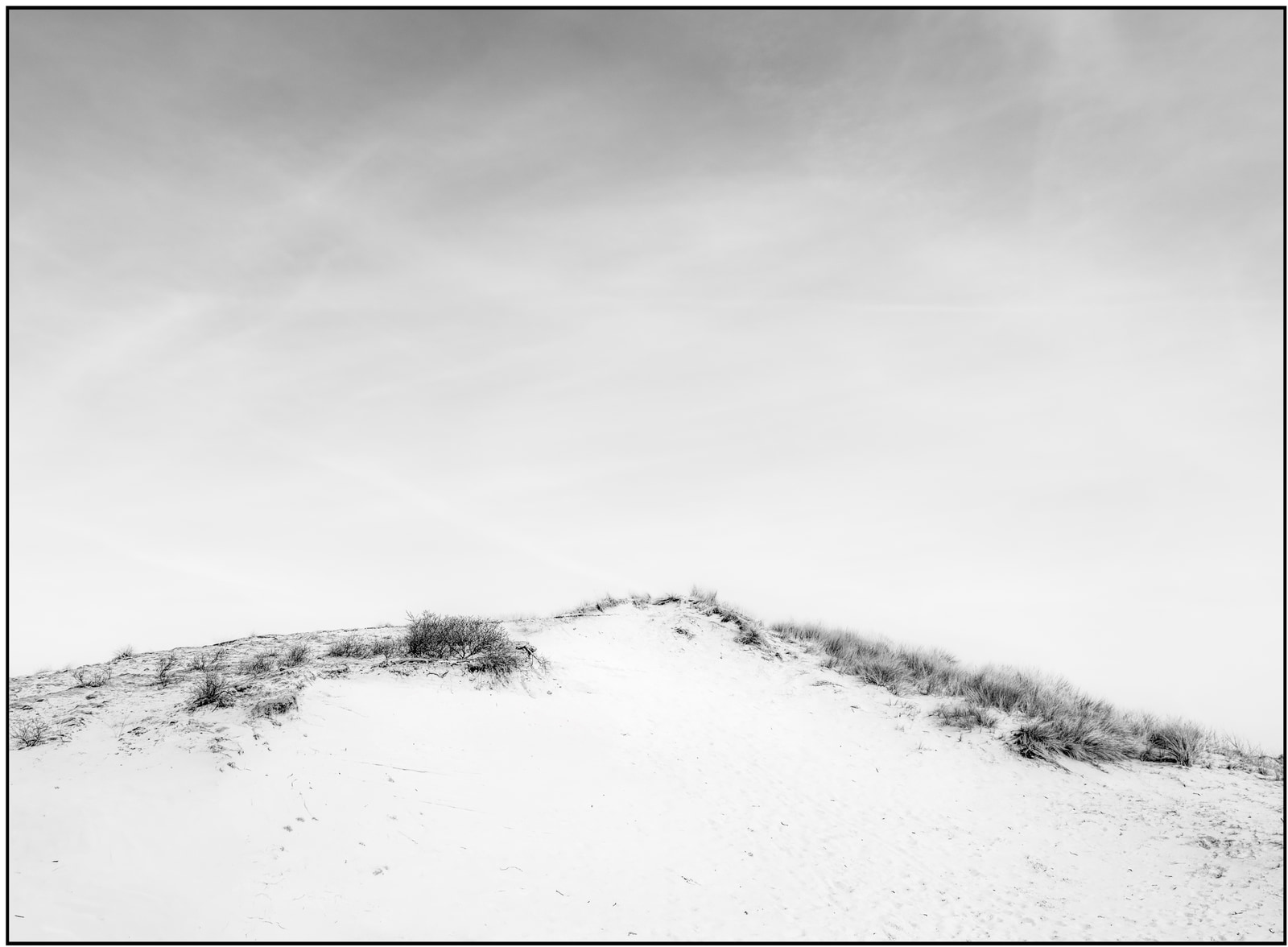A sandy dune rises under a cloudy sky, with sparse vegetation dotting its surface.