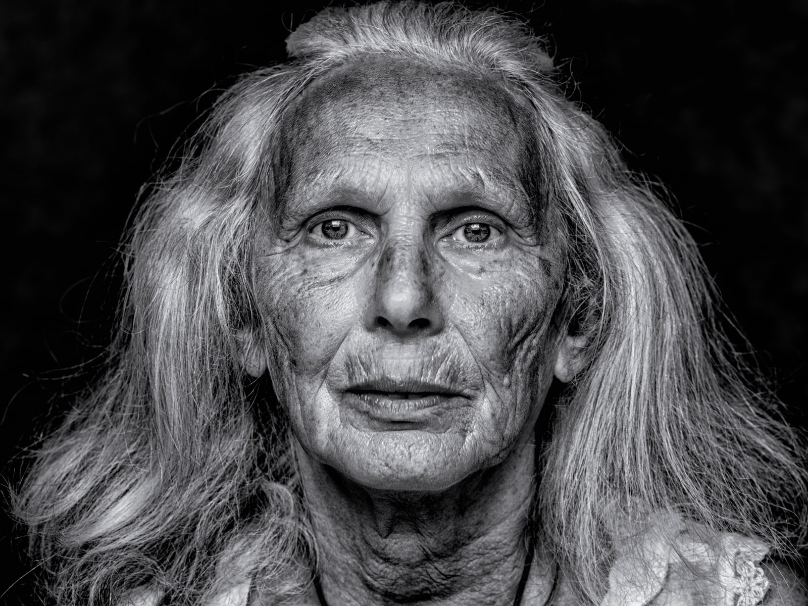 An elderly person with long hair and a serious expression is shown in a close-up portrait.