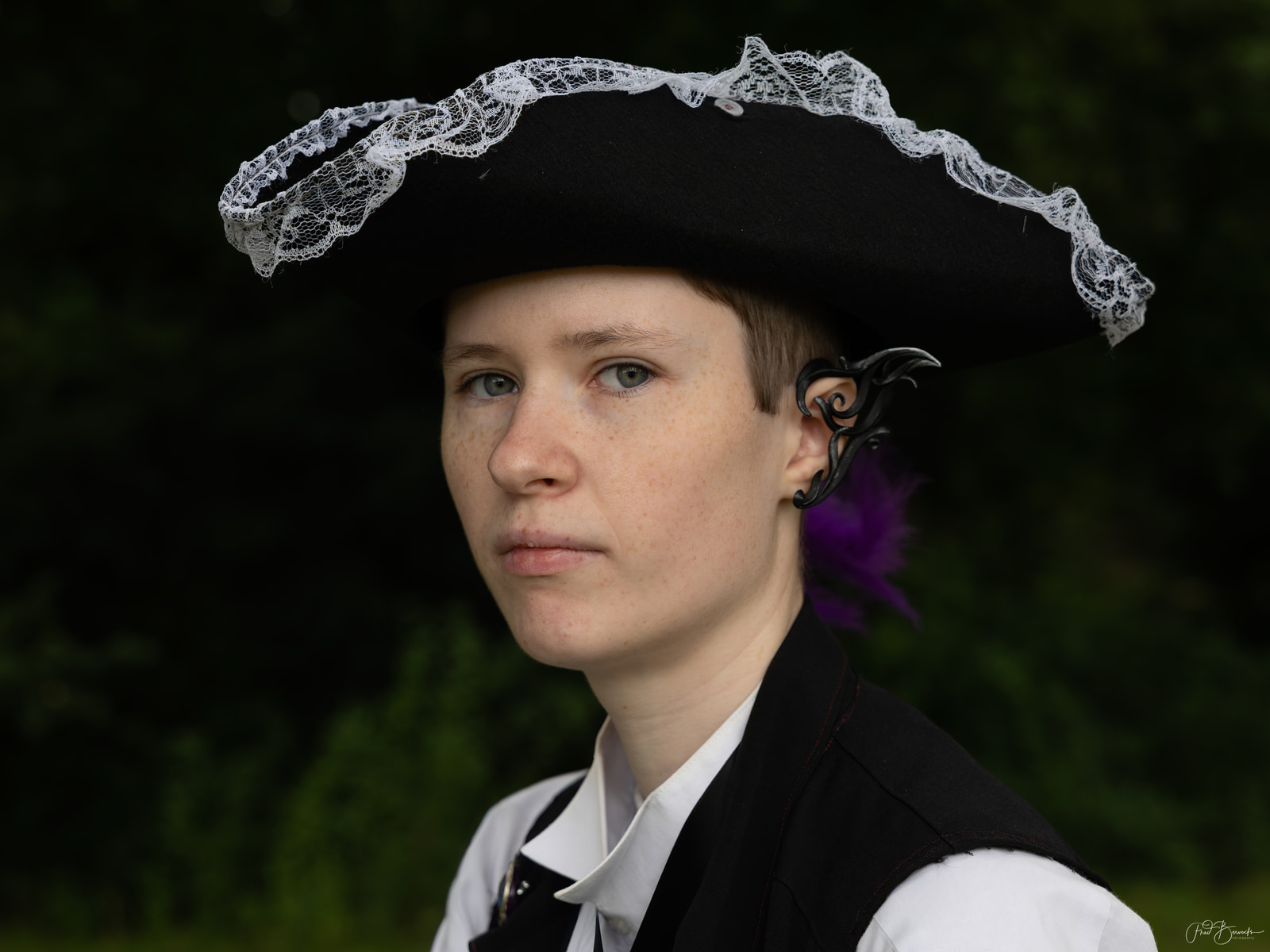 A person in period costume with a tricorn hat and lace trim, wearing an ornate earring and a black vest over a white shirt.