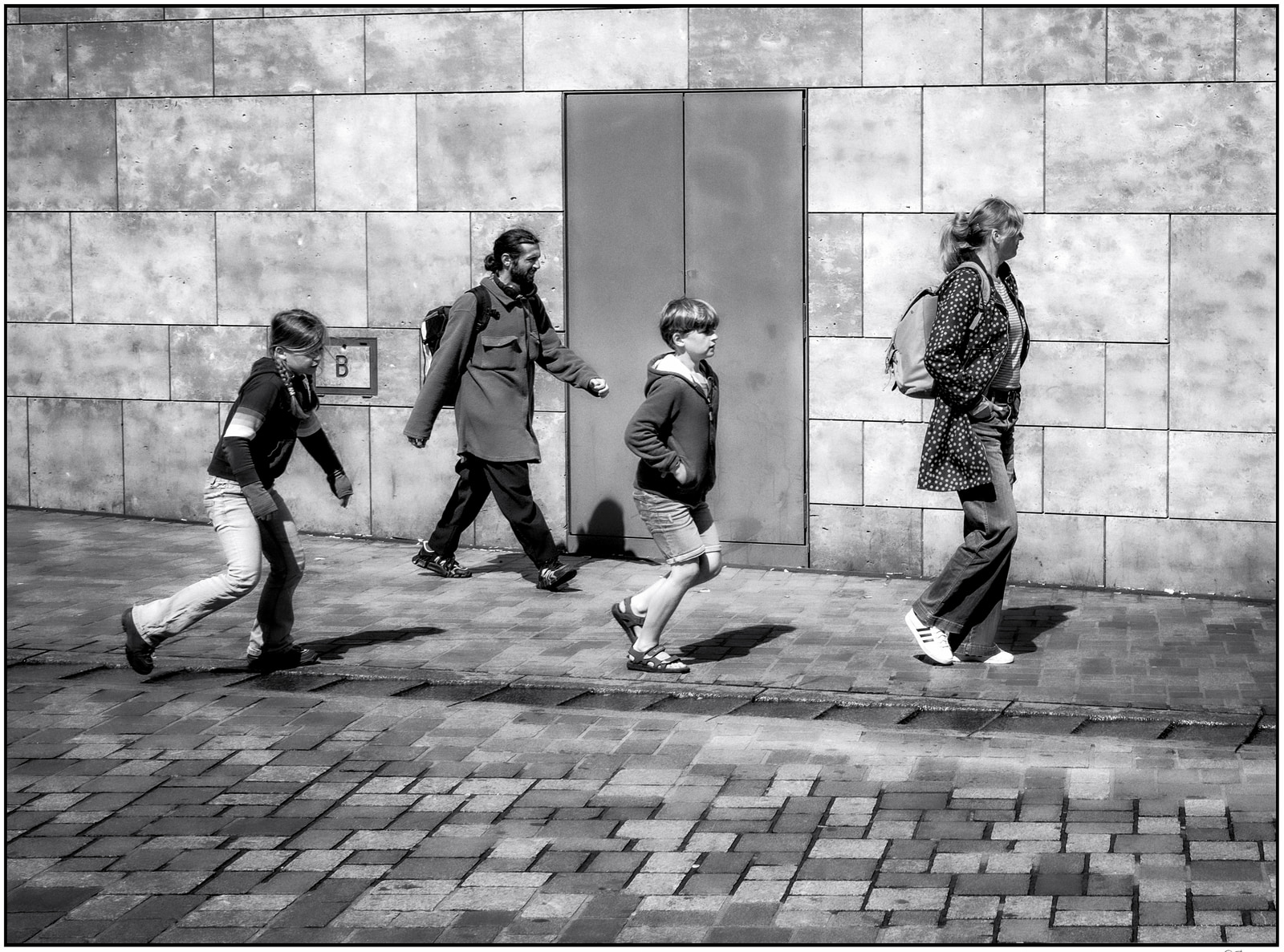 Four people walk in a line along a paved sidewalk beside a tall concrete wall.