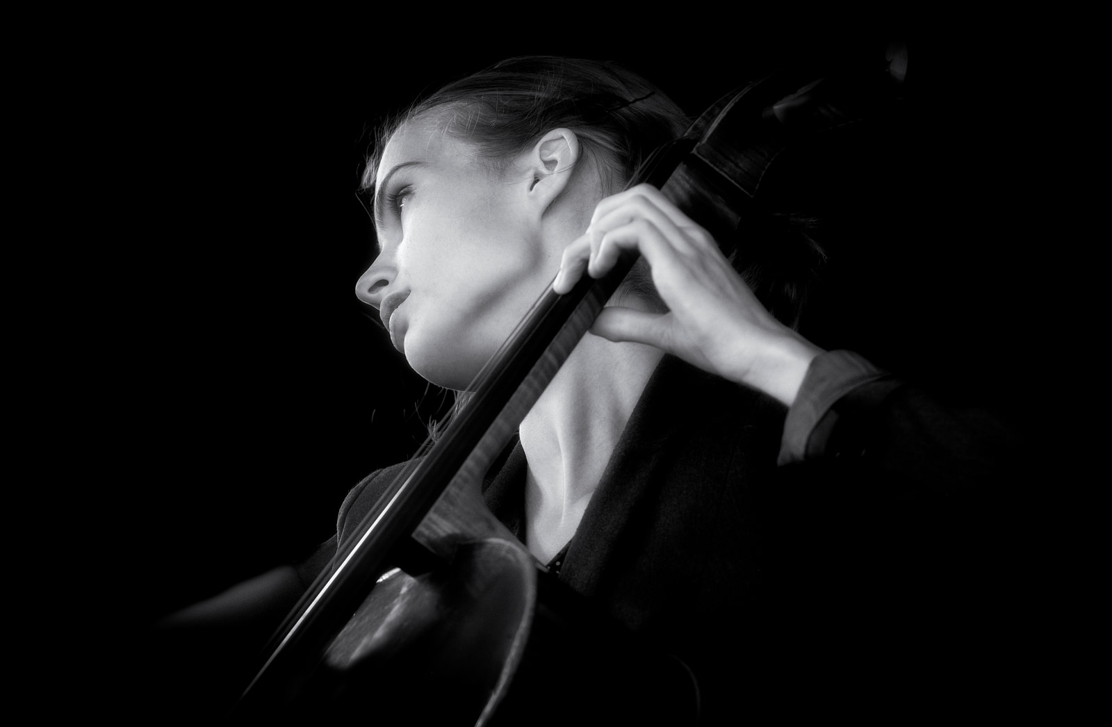 A person plays a cello in a dark setting, their face and instrument highlighted against the shadows.