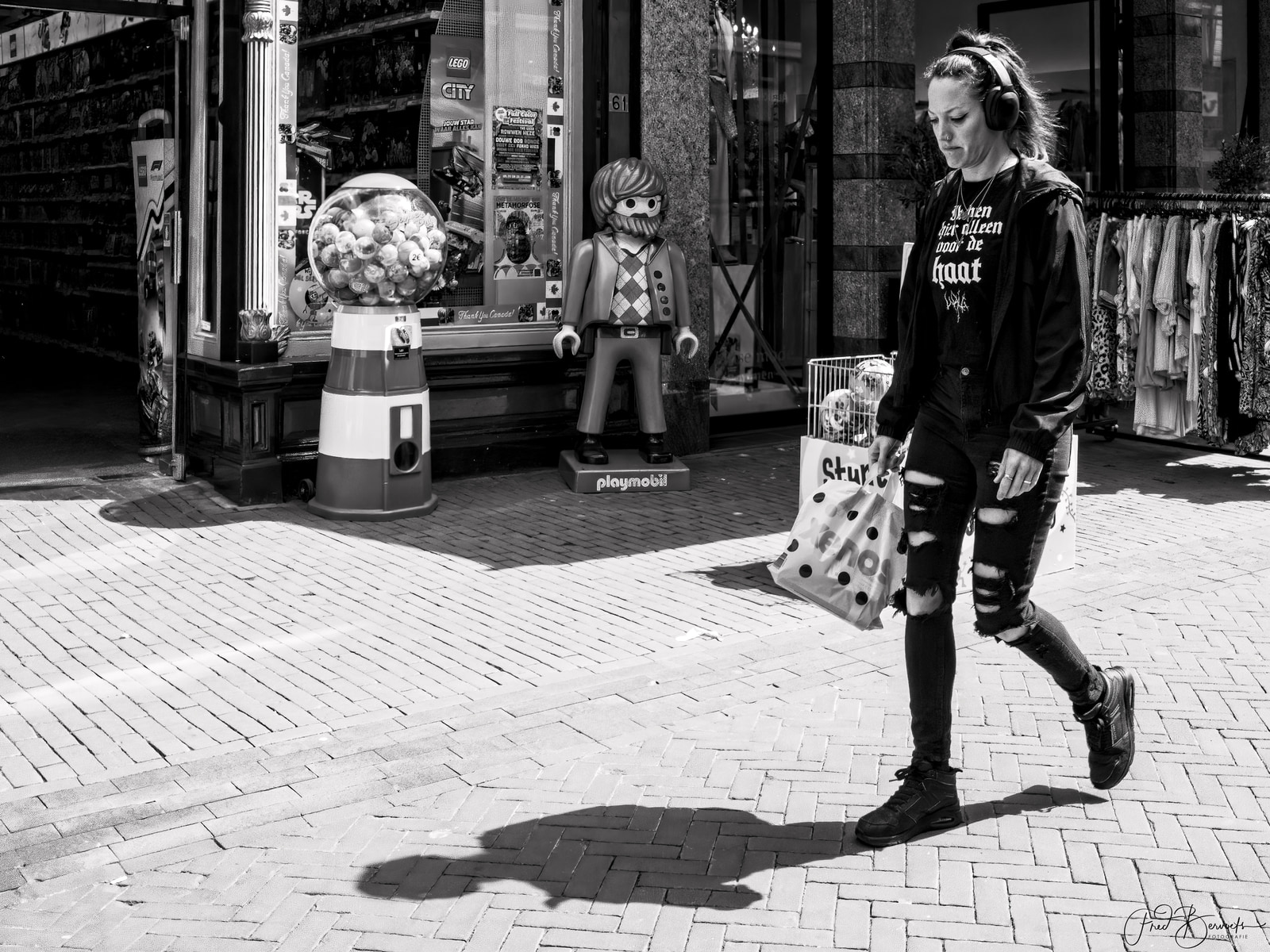 A person wearing headphones walks past a storefront with a large Lego display and a gumball machine, carrying a shopping bag.