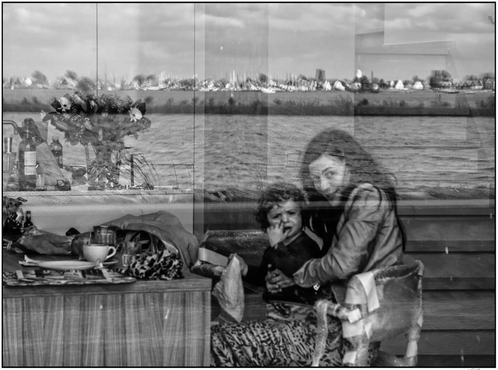 A woman cradles a child in her lap, both seated near a table with cups and a vase of flowers, with a body of water and distant cityscape visible through a large window behind them.