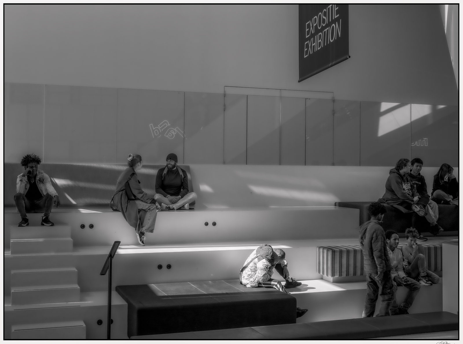 Several people sit and stand on a tiered platform inside a modern gallery space, with one person crouching near the steps.