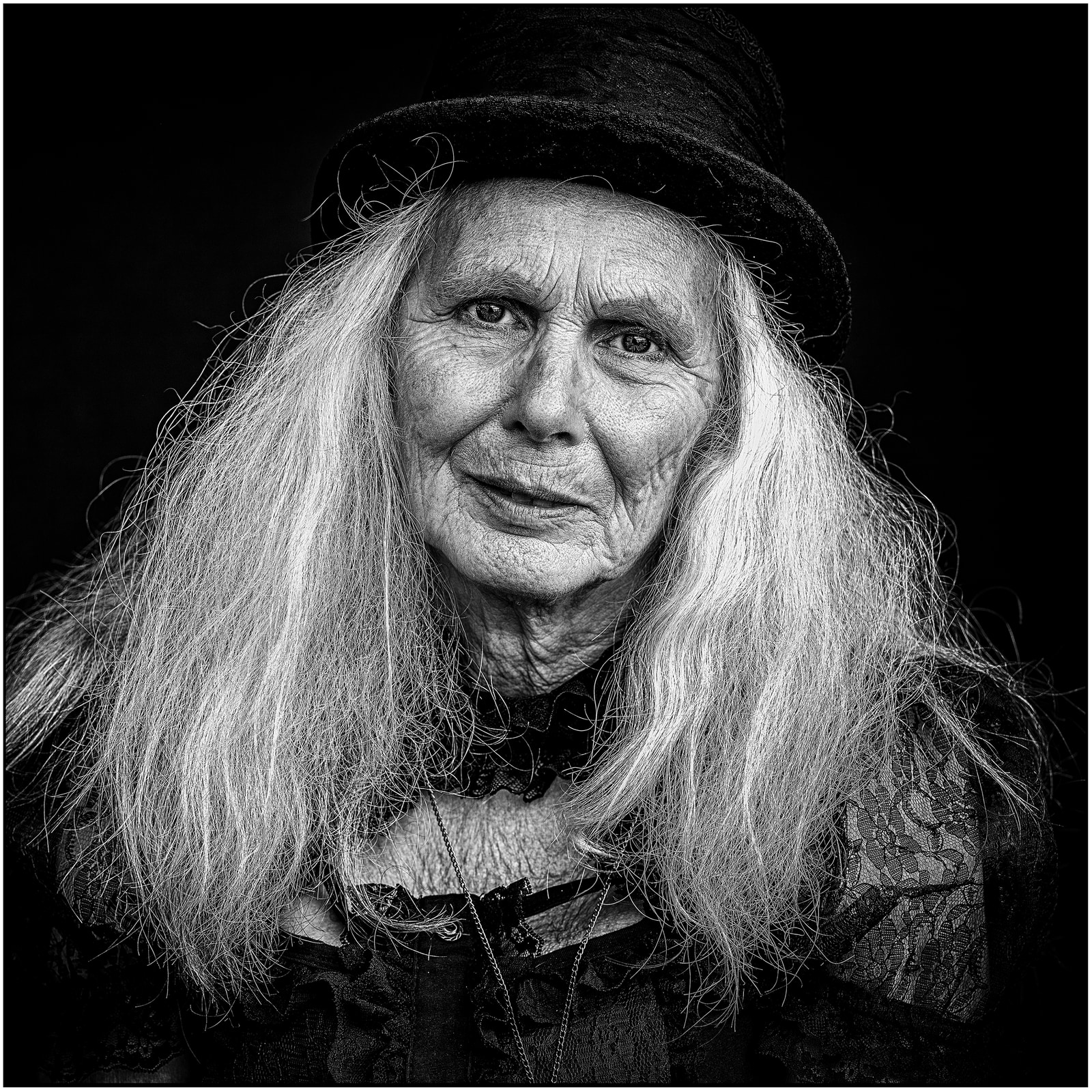 An elderly person with long hair and a hat gazes directly at the camera against a dark background.