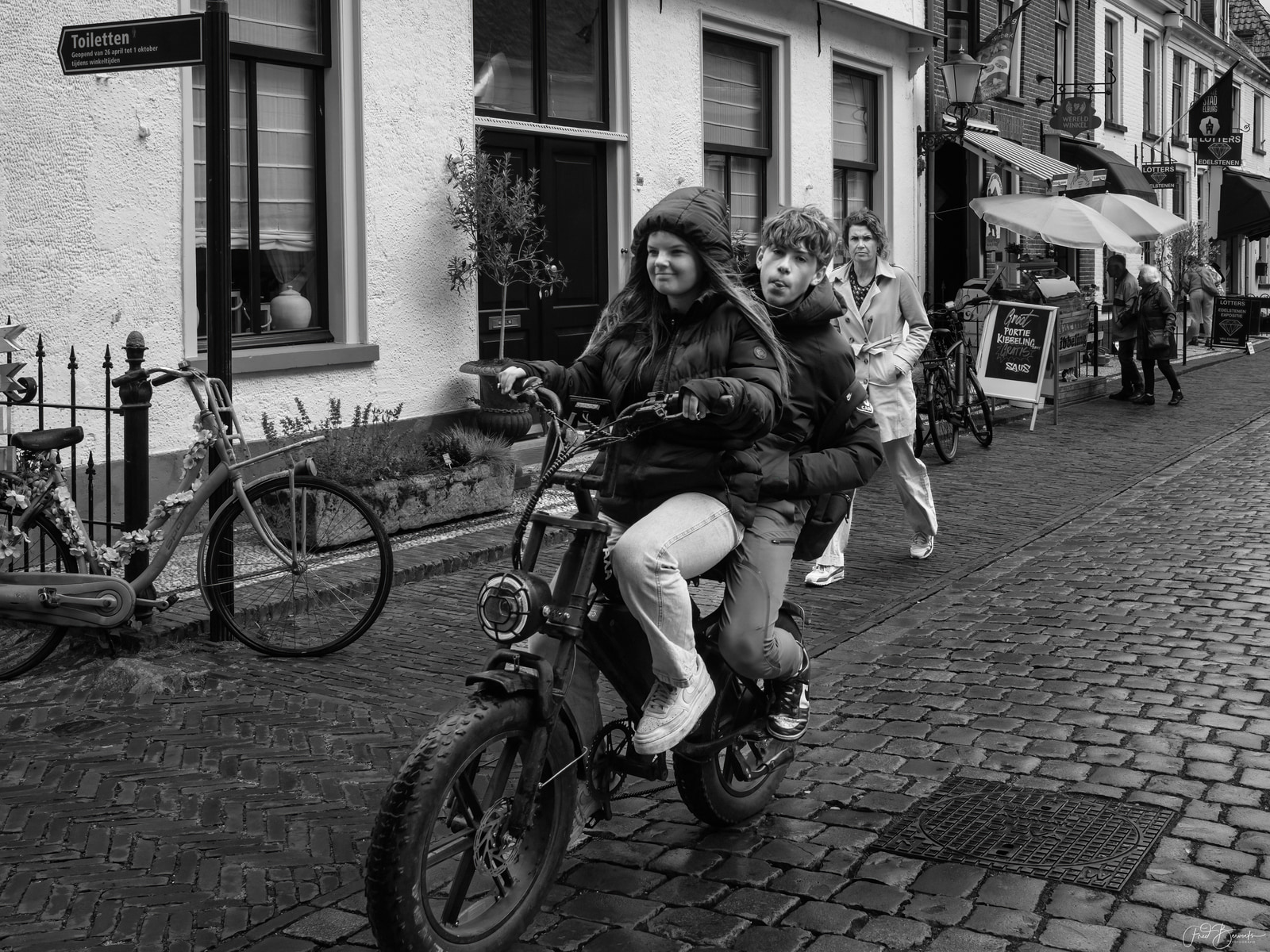 A young person rides a small motorcycle down a cobblestone street, with another person seated behind, passing storefronts and parked bicycles.