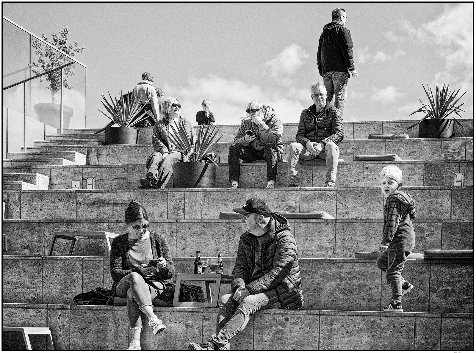 Several people sit and stand on concrete steps outdoors, some using phones, with potted plants nearby.