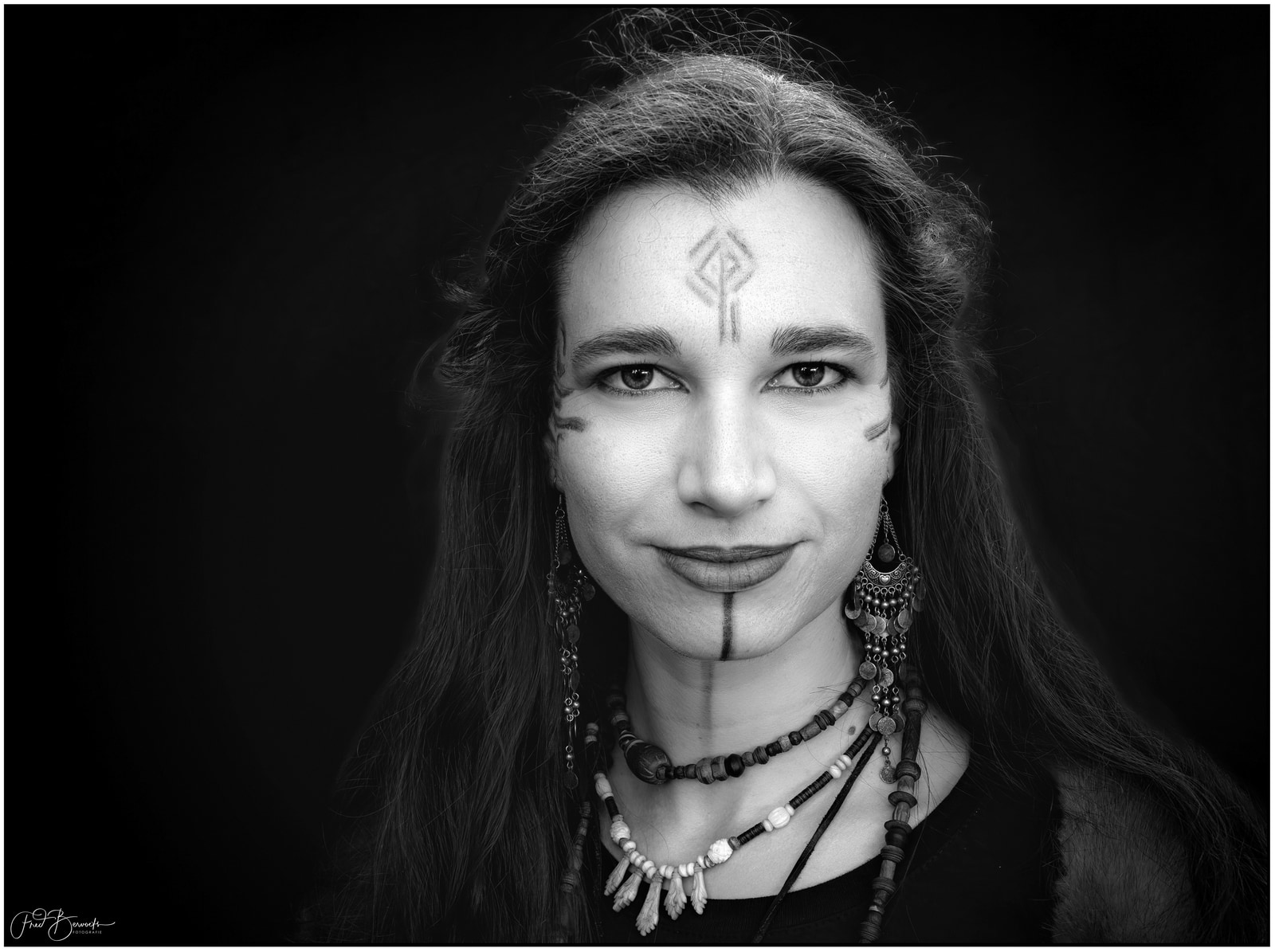 A person with long hair and intricate facial markings, wearing layered beaded necklaces, is shown in a close-up portrait against a dark background.