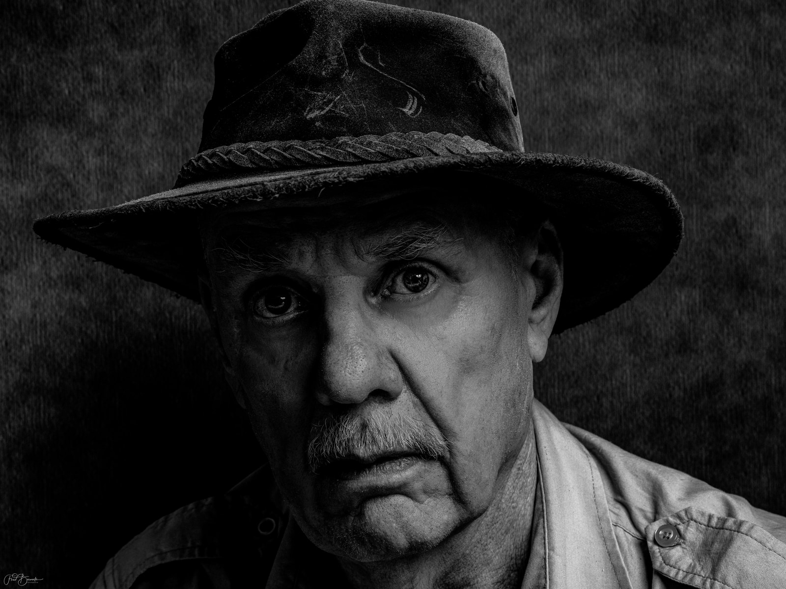 An older man with a mustache and a hat looks directly at the camera.