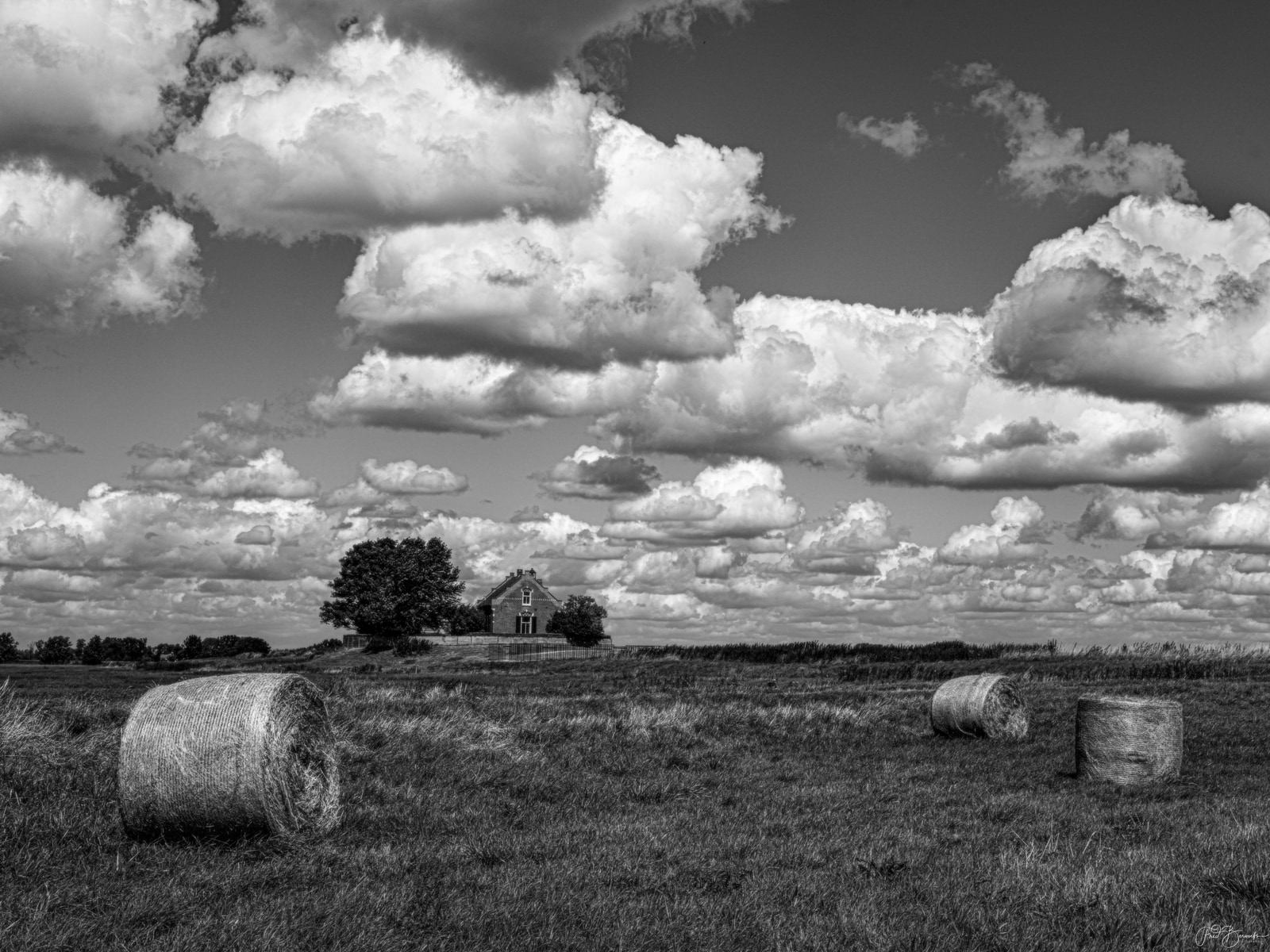 Bales of hay lie in a field beneath a dramatic sky, with a solitary house visible in the distance.