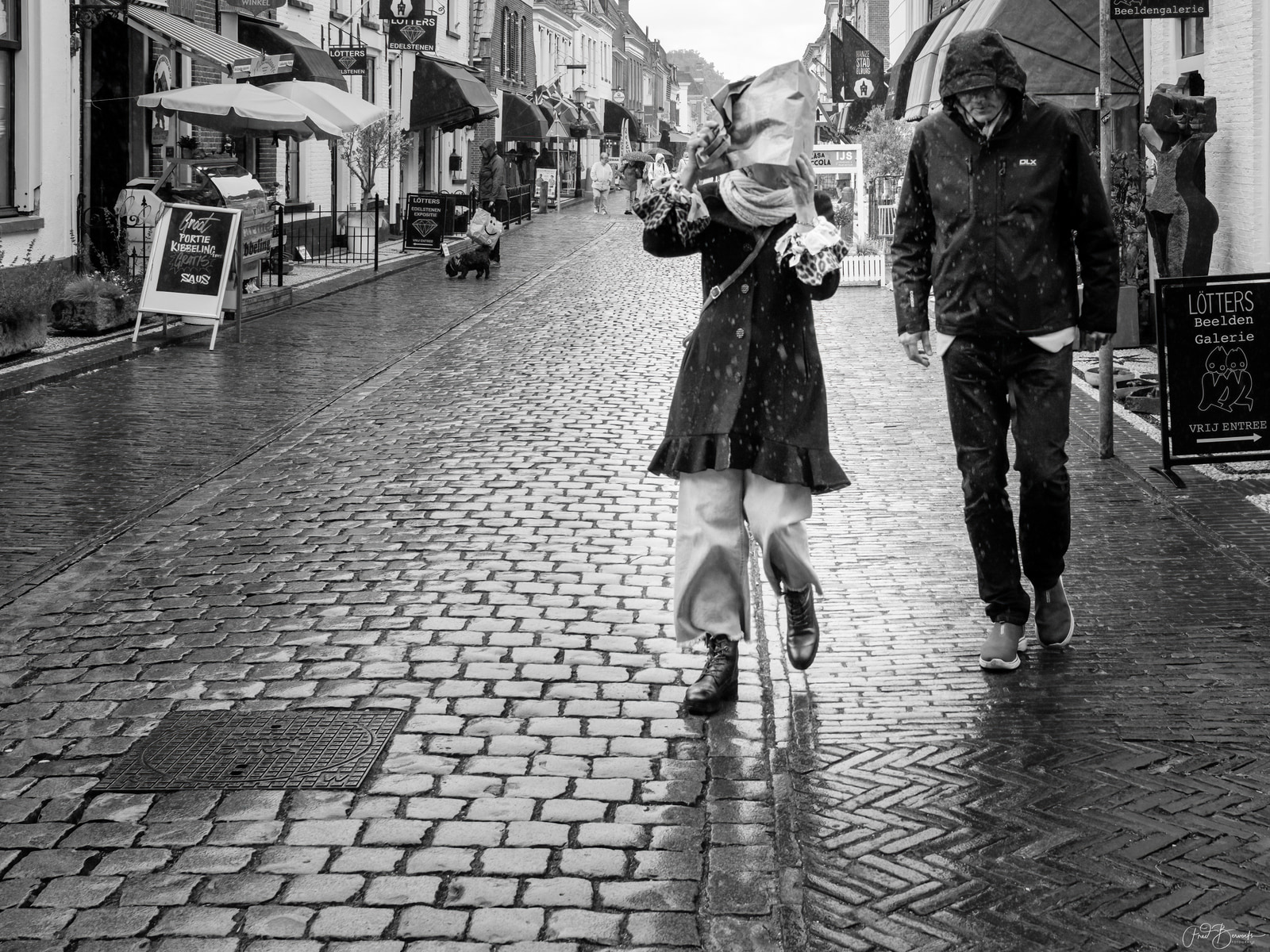 Two people walk on a wet cobblestone street in a town square, one carrying an umbrella and a bag, while the other wears a hooded jacket.