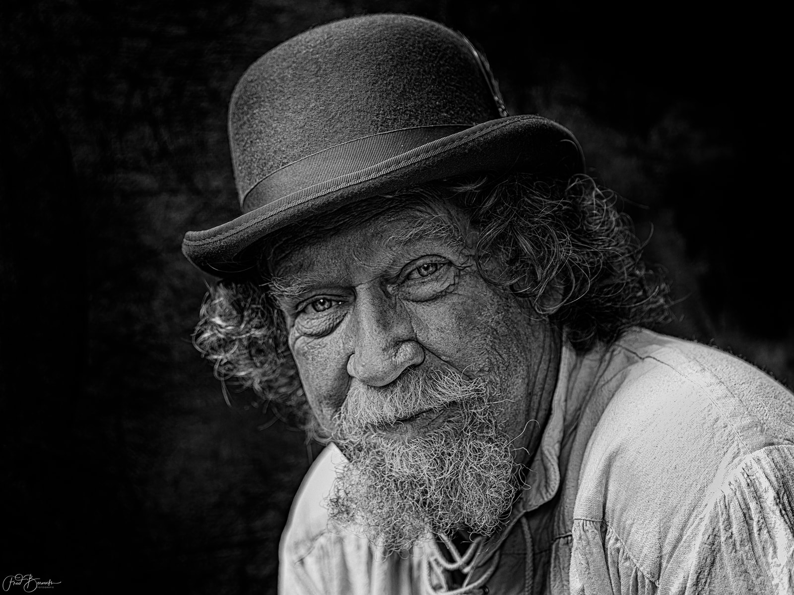An older man with a long beard and mustache, wearing a hat, gazes directly at the camera.