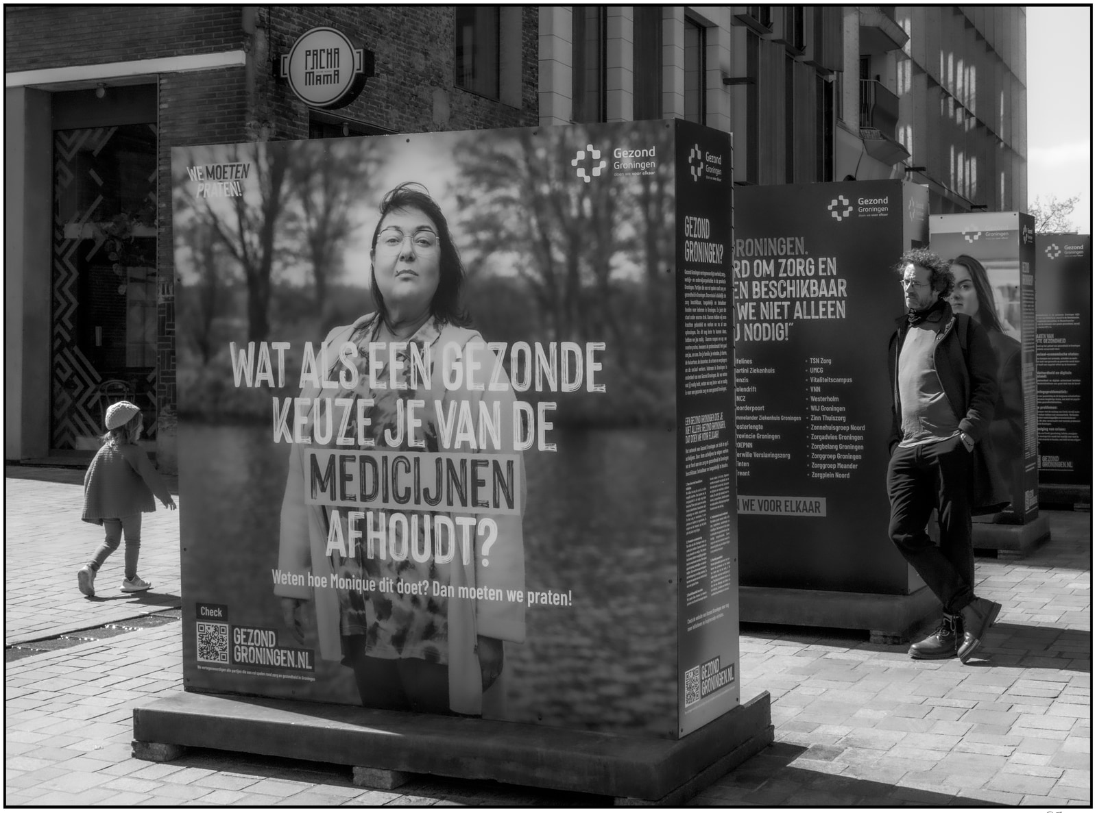 A black-and-white street scene shows a large public display board with text in Dutch, while two people walk past on the sidewalk.