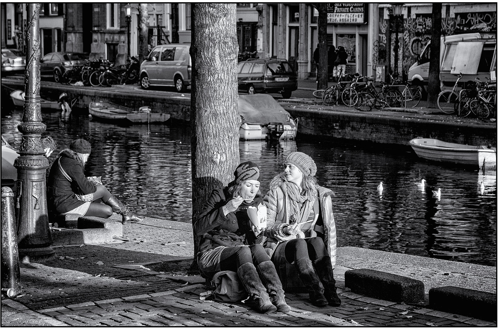 Three people sit on a stone ledge by a canal, eating and talking, with boats and bicycles along the waterfront.