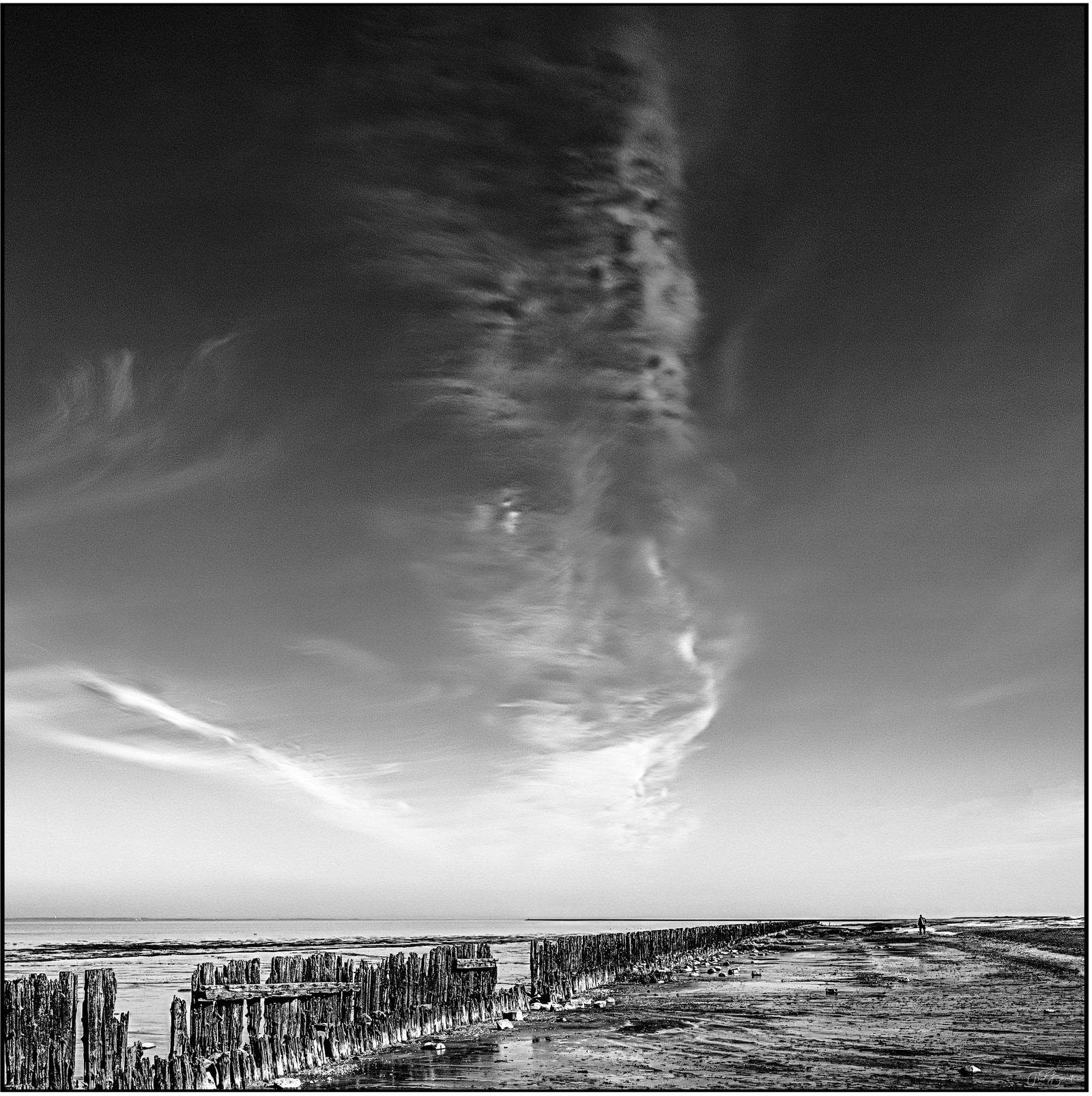 A lone figure stands on a sandy beach beside weathered wooden posts, with a dramatic, swirling cloud formation dominating the sky above the calm sea.