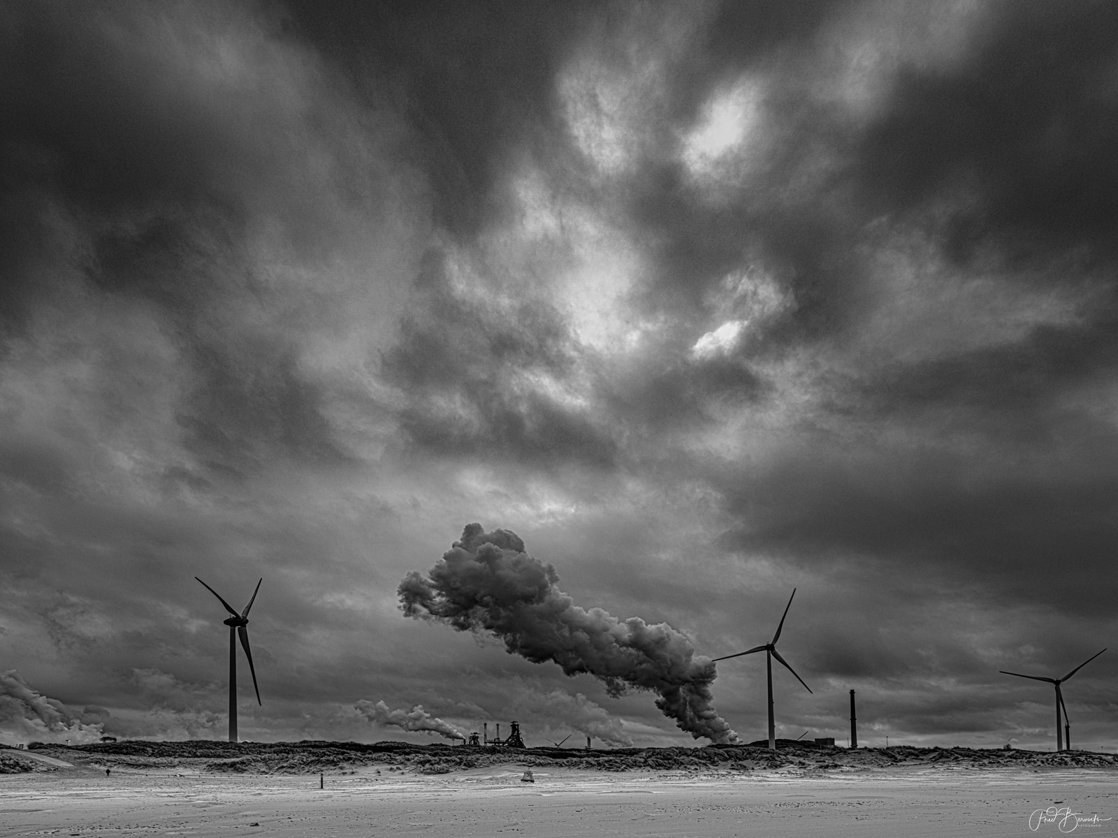 Wind turbines stand against a dramatic, stormy sky with dark, heavy clouds and a faint light breaking through.