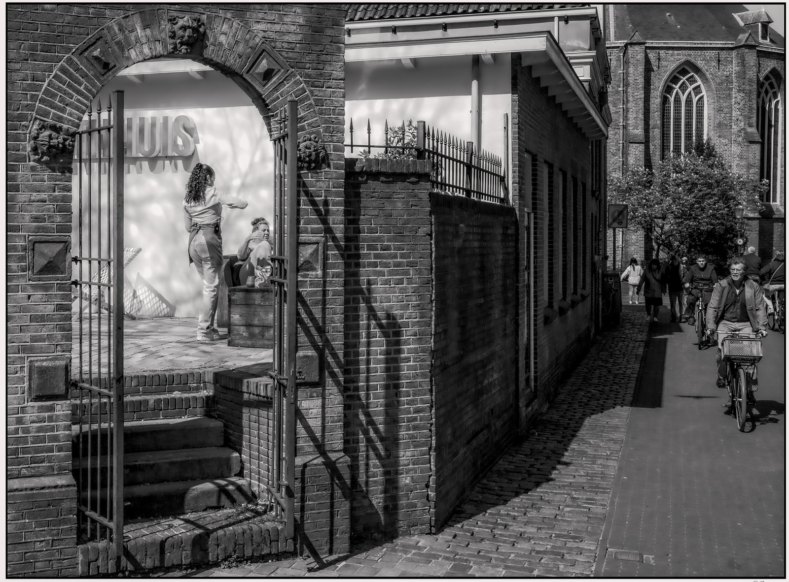 A black-and-white street scene shows a brick archway with a person standing on steps, while a cyclist passes by on the cobblestone road beside historic buildings.