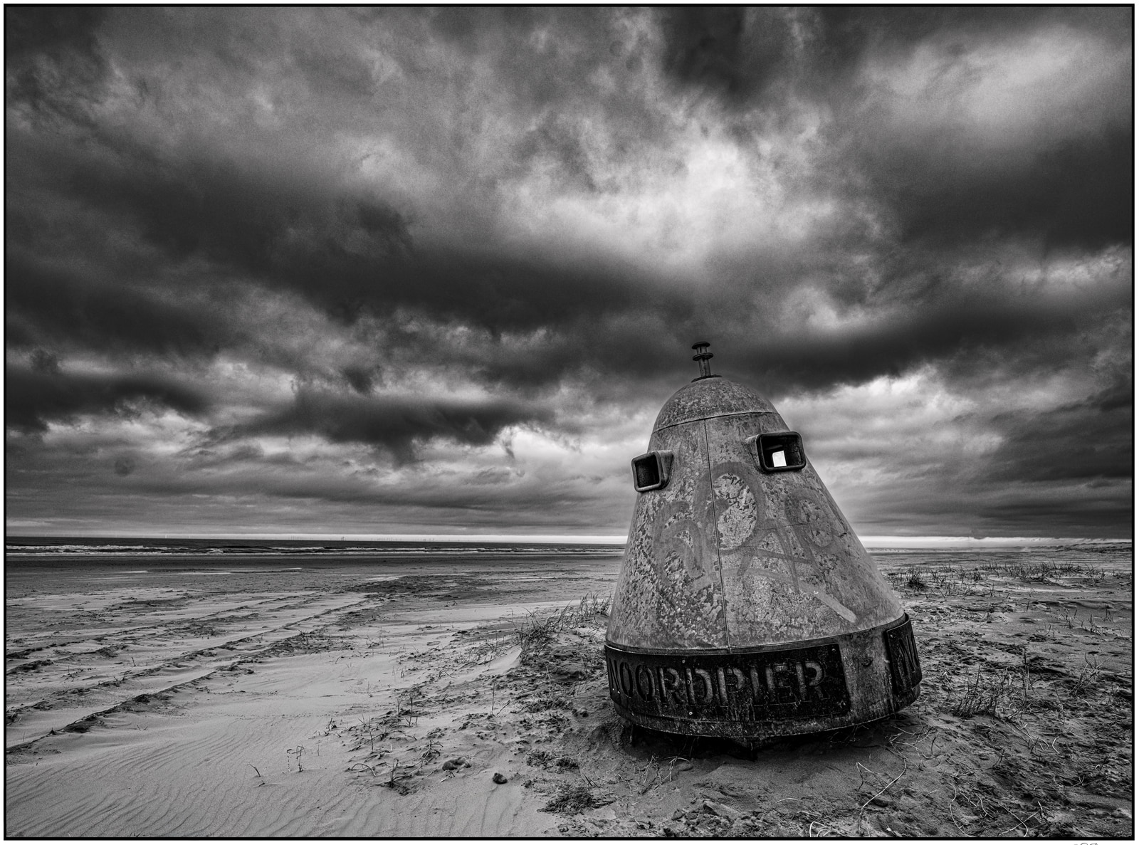 A weathered, cylindrical concrete structure stands on a barren, sandy landscape under a dramatic, overcast sky.