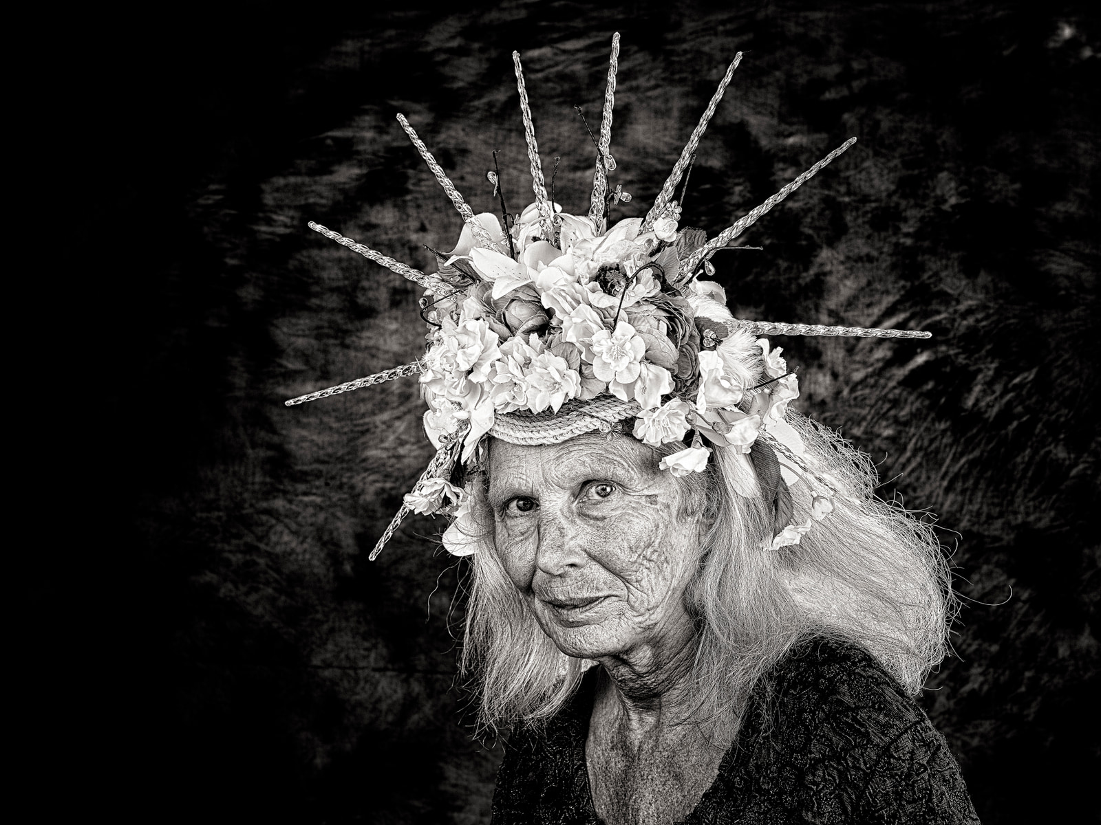 An elderly person smiles while wearing a decorative headpiece with flowers and thin branches.