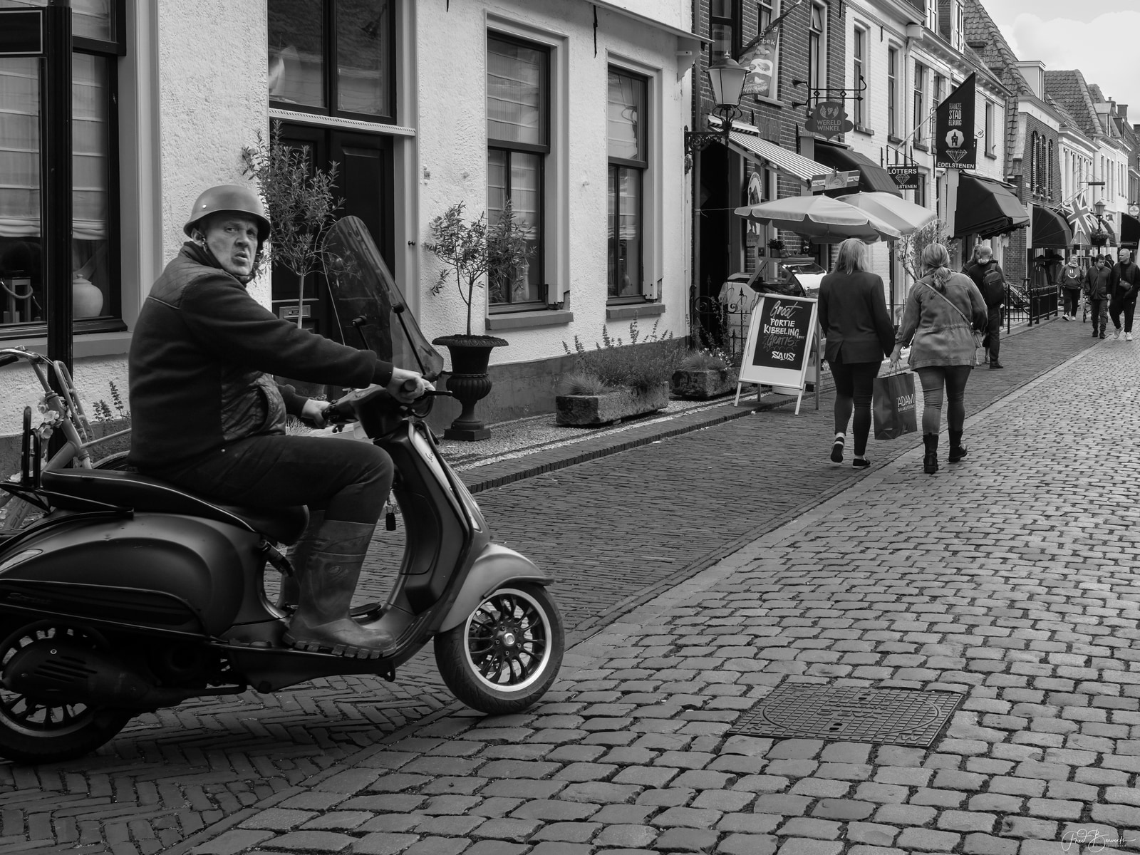 A man in a helmet rides a scooter down a cobblestone street past shops and pedestrians.