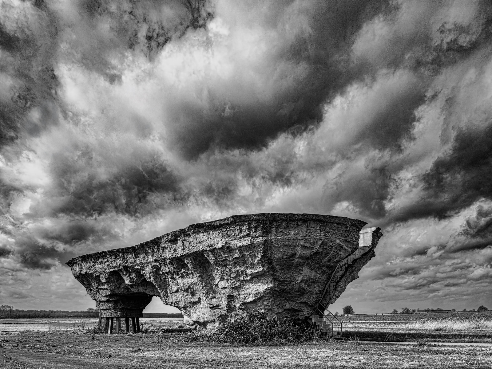 A large, weathered concrete structure with a curved roof stands in an open field under dramatic, brooding clouds.