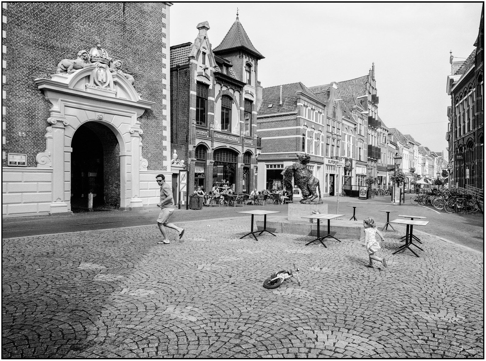 A man and a child walk through a cobblestone square beside a stone archway and ornate buildings, with bicycles and outdoor tables nearby.