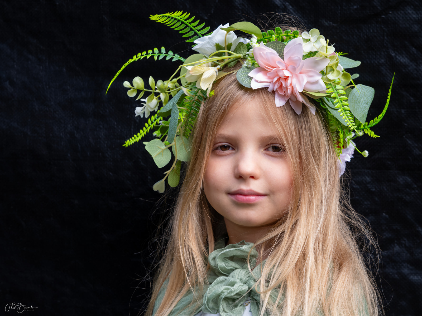 A young girl with long blonde hair wears a floral headband featuring white and pink flowers against a dark background.