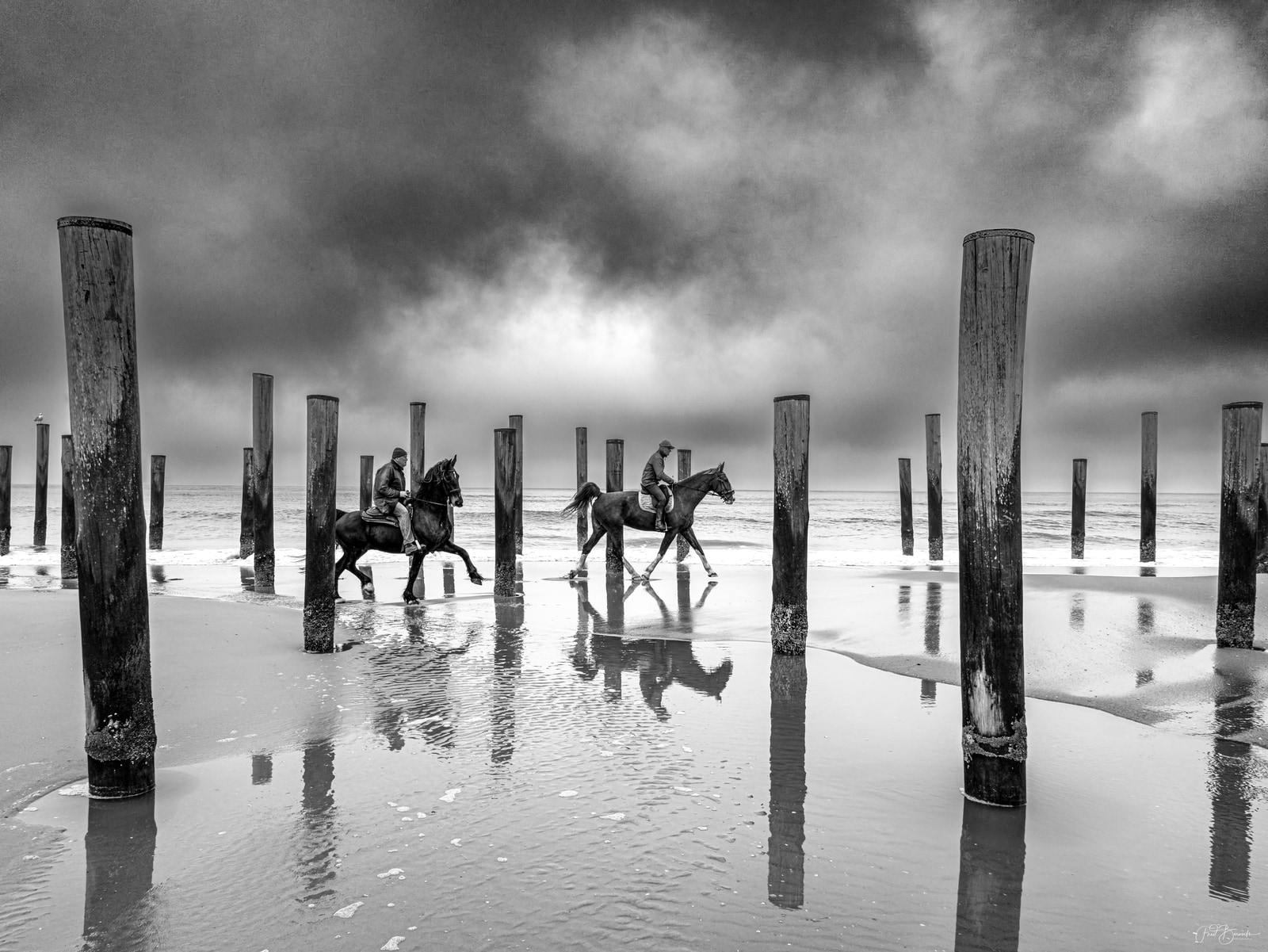 Two riders on horseback cross a wet beach through a row of wooden posts under a stormy sky.