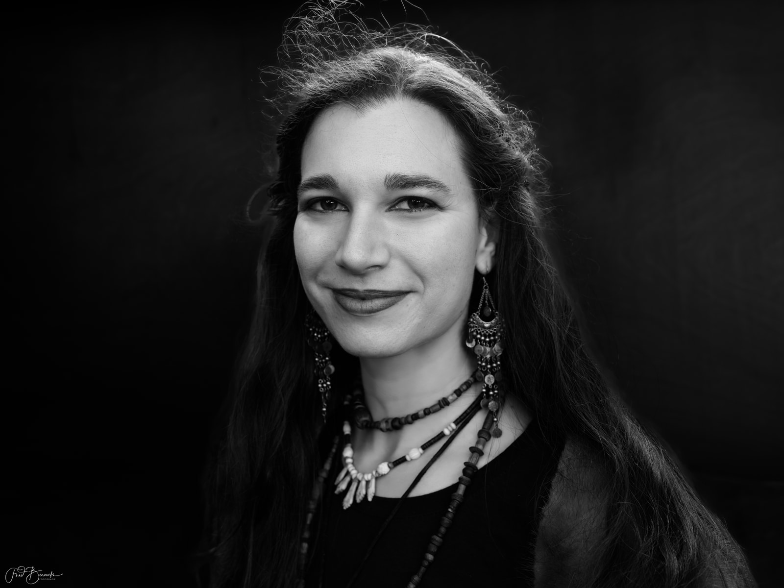 A woman with long hair and earrings smiles, wearing layered beaded necklaces against a dark background.