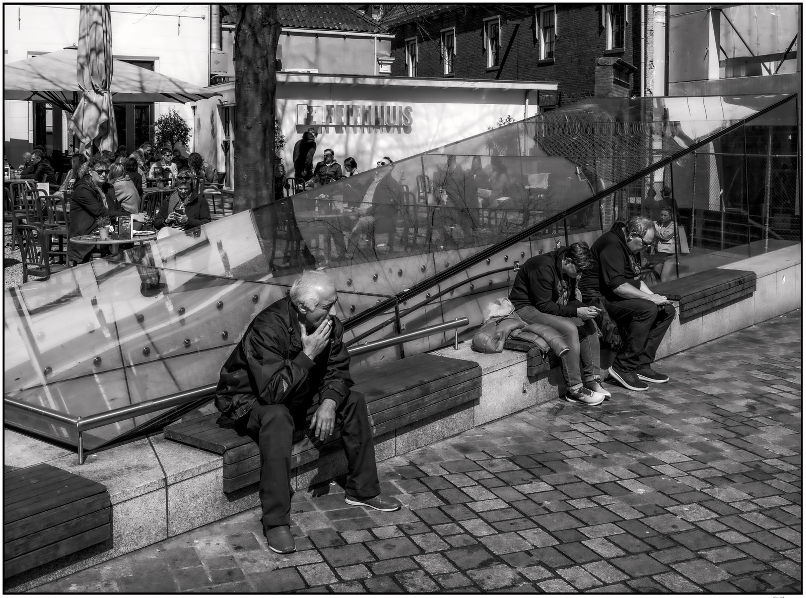 People sit on benches and at tables in an outdoor café area, with a glass barrier and a tree in the foreground.