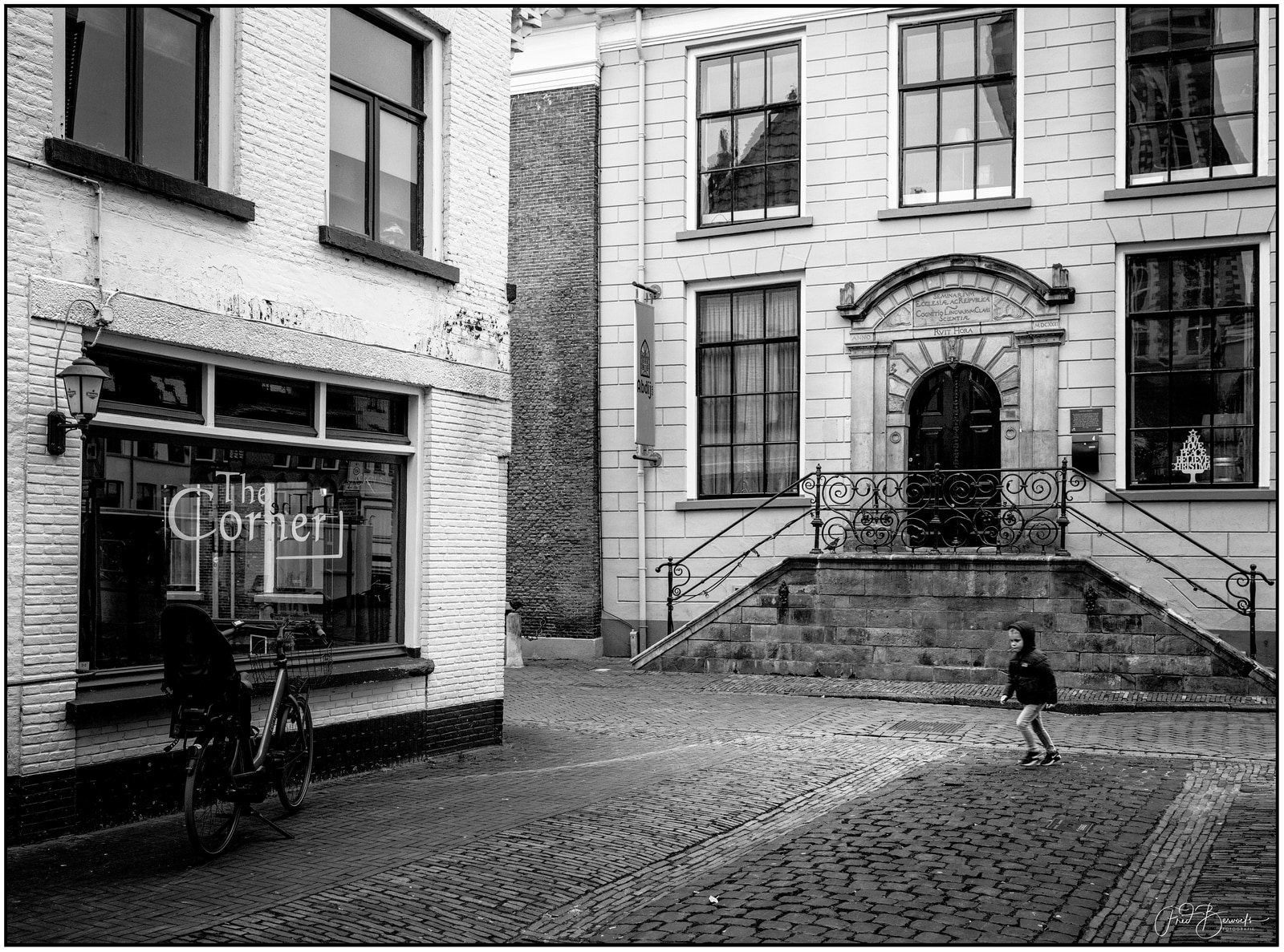 A child walks on a cobblestone street beside a building with a staircase and ornate doorway, while a bicycle leans against the wall.