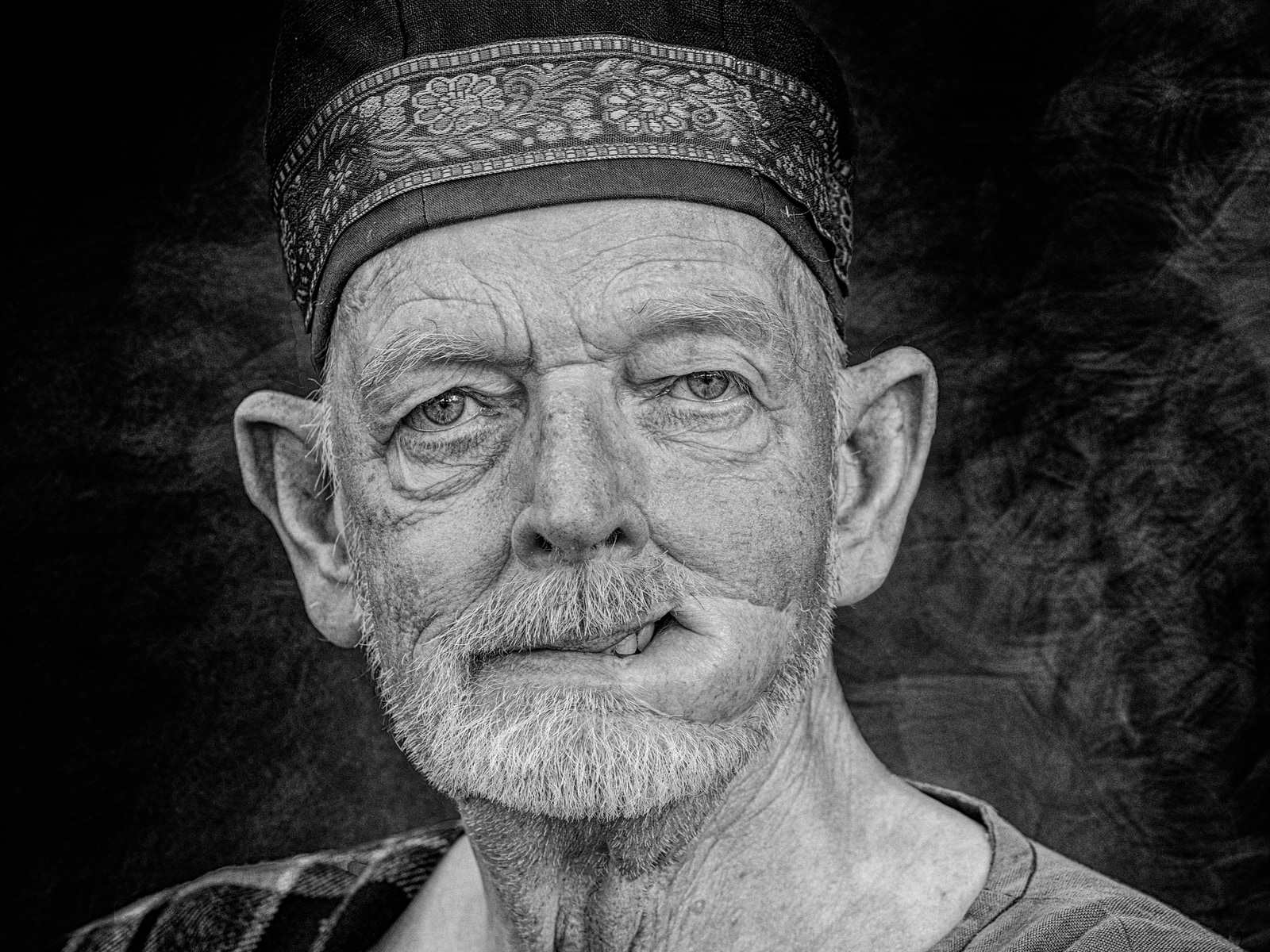 An elderly man with a beard and mustache, wearing a patterned hat, looks directly at the camera.