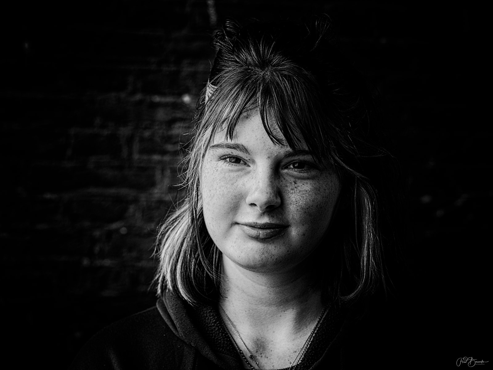 A person with short hair and a gentle smile is captured in a close-up portrait against a dark, out-of-focus background.