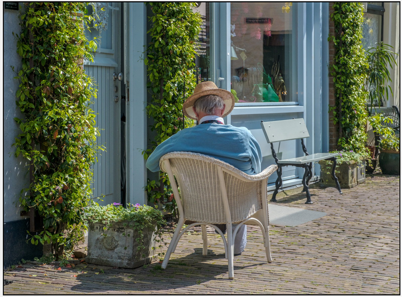 An older man in a straw hat sits on a wicker chair outside a building, with potted plants and a bench nearby.