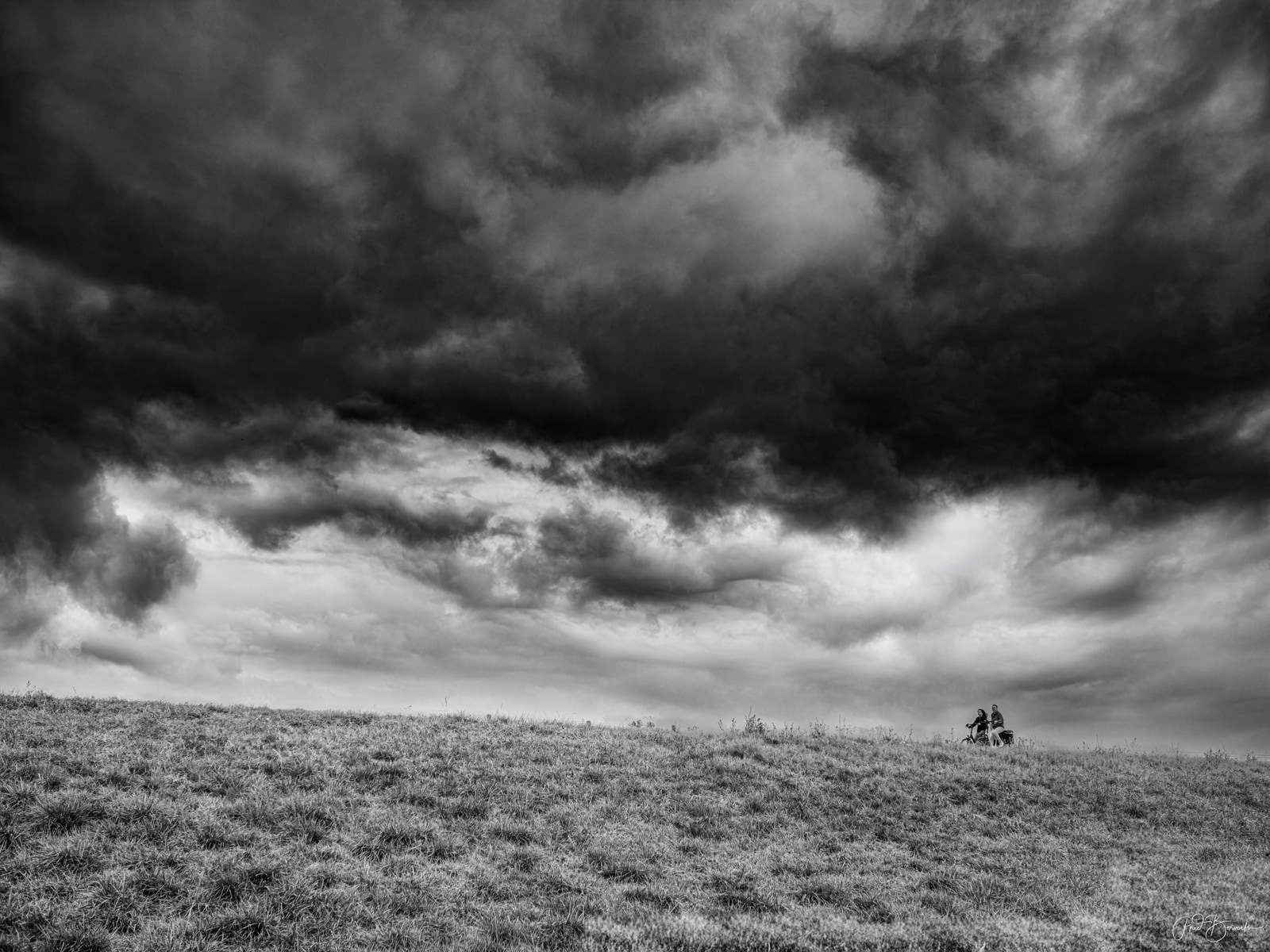 Two cyclists ride across a grassy hill under a dramatic, brooding sky.