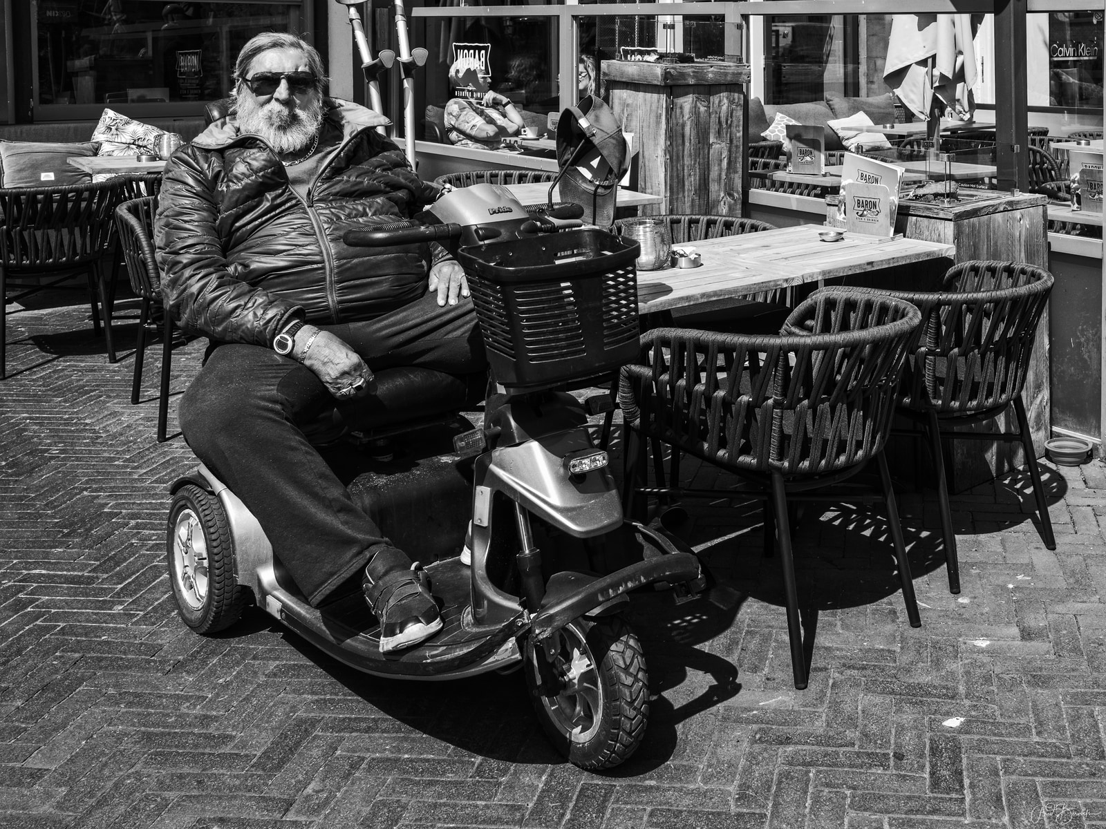 An older man sits on a three-wheeled mobility scooter at an outdoor café table.