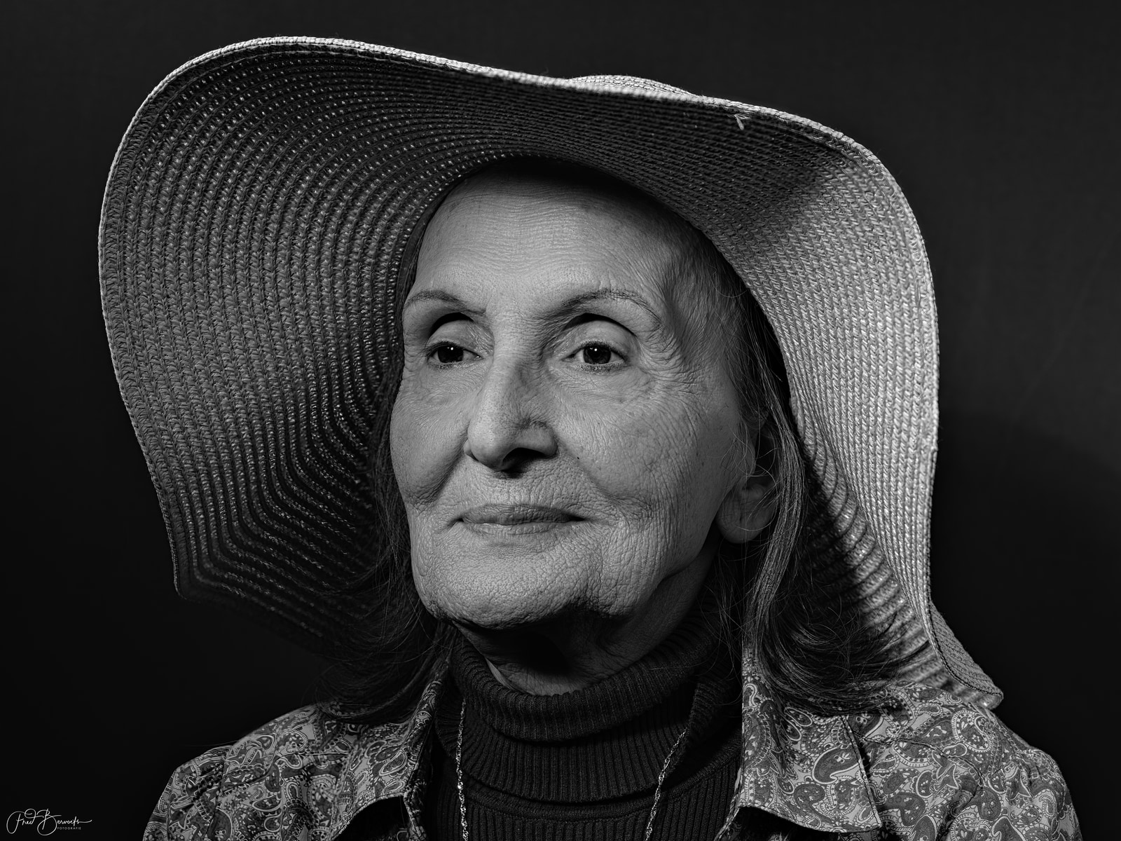 An elderly woman with long hair gazes directly at the camera, wearing a wide-brimmed hat and a patterned shawl.