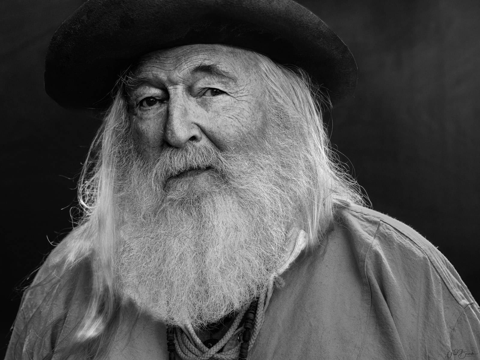 An elderly man with long white hair and beard, wearing a dark hat, gazes directly at the camera.