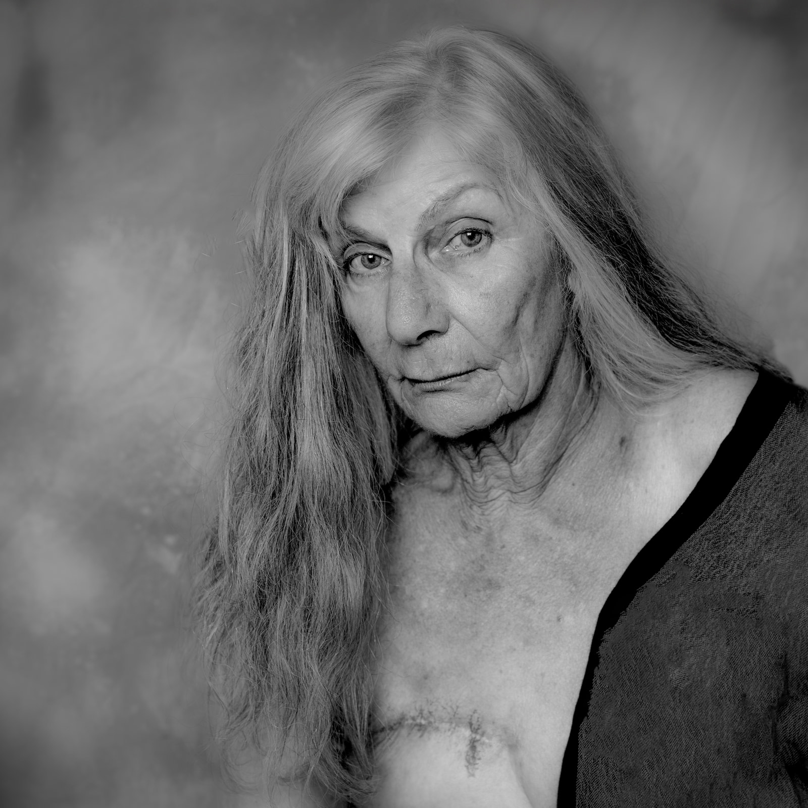A close-up portrait of an older woman with long hair, wearing a dark top, against a softly blurred background.