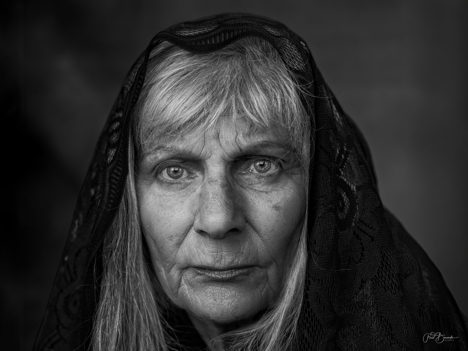 An elderly woman with light hair and a patterned headscarf gazes directly at the camera.