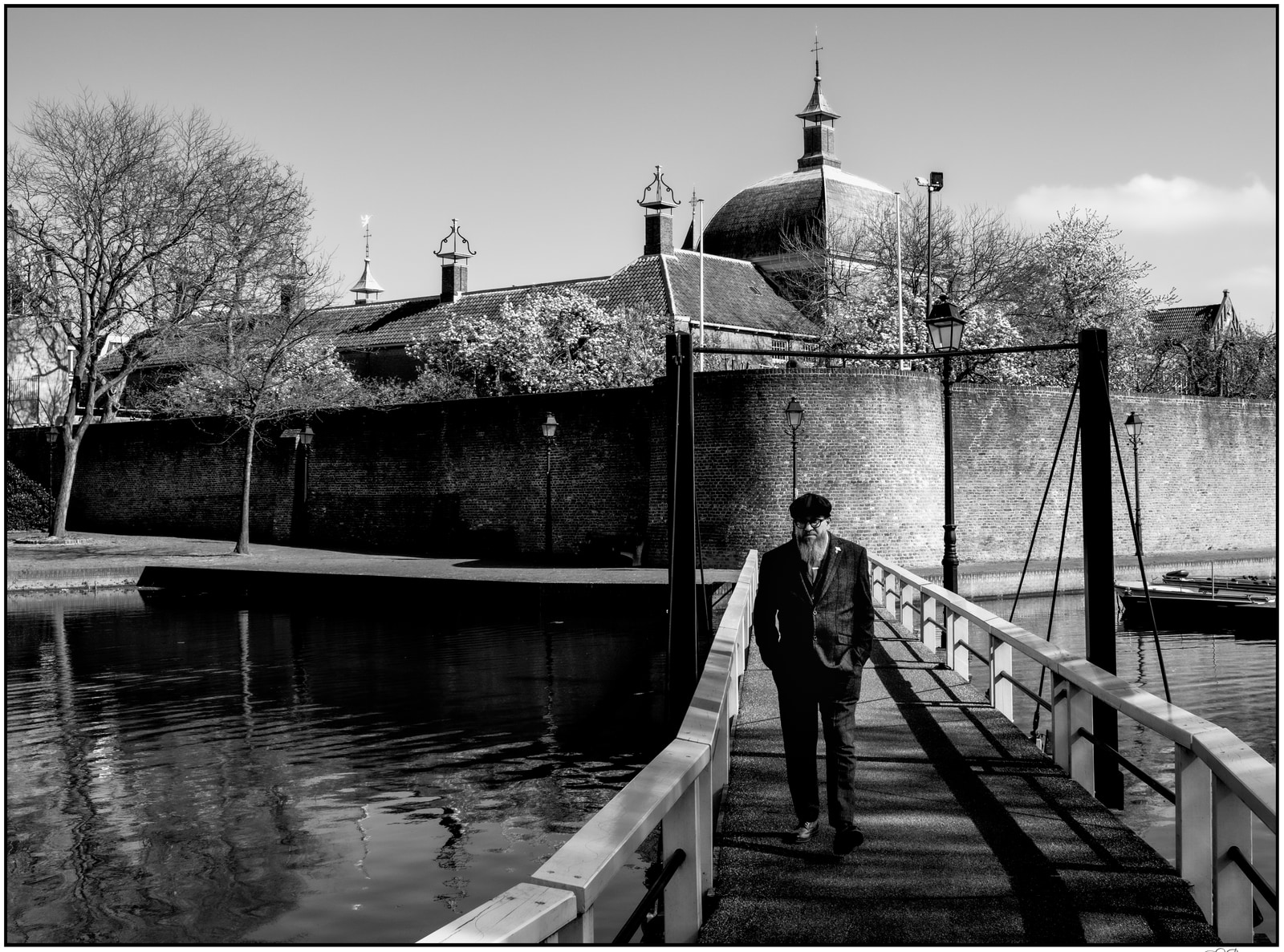 A man in a suit walks along a narrow bridge beside a canal, with a domed building and stone walls in the background.