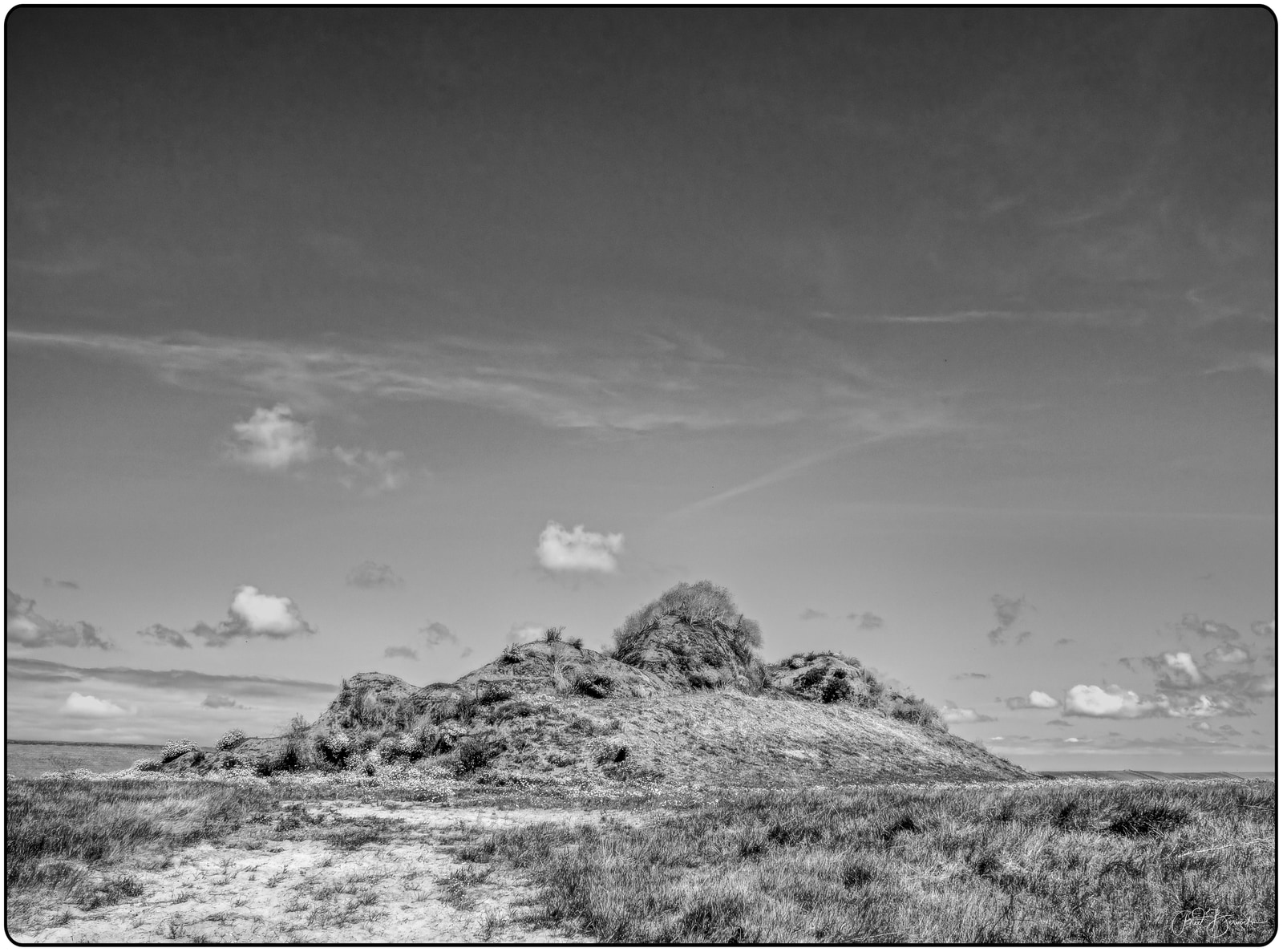 A lone hill rises from flat grassland under a dramatic sky with scattered clouds.