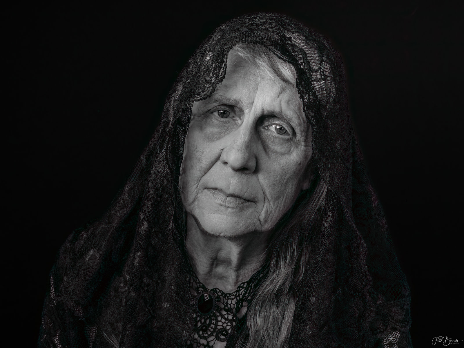 An elderly person with long hair and a lace head covering, facing the camera against a dark background.