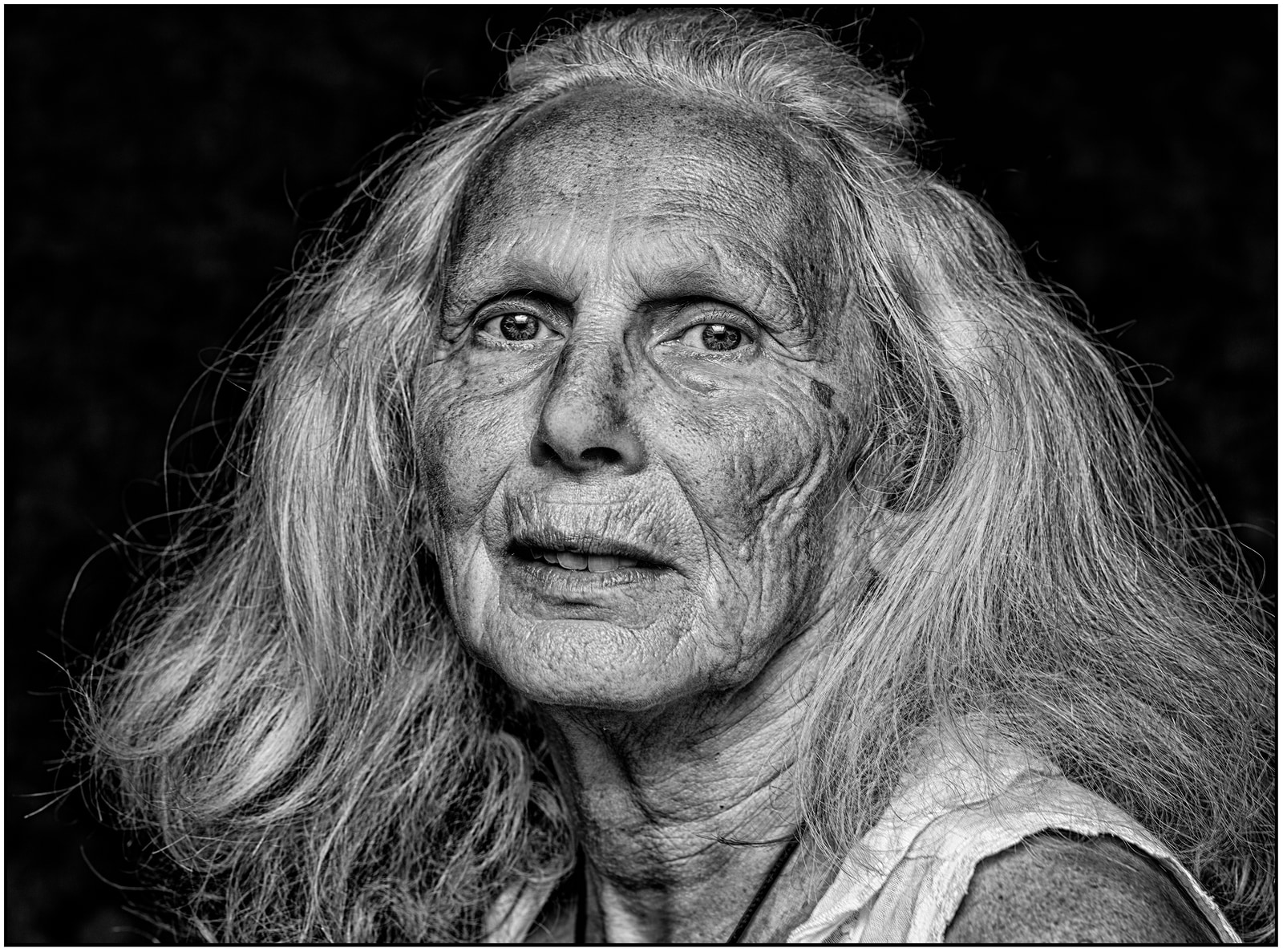 An elderly person with long hair gazes directly at the camera against a dark background.