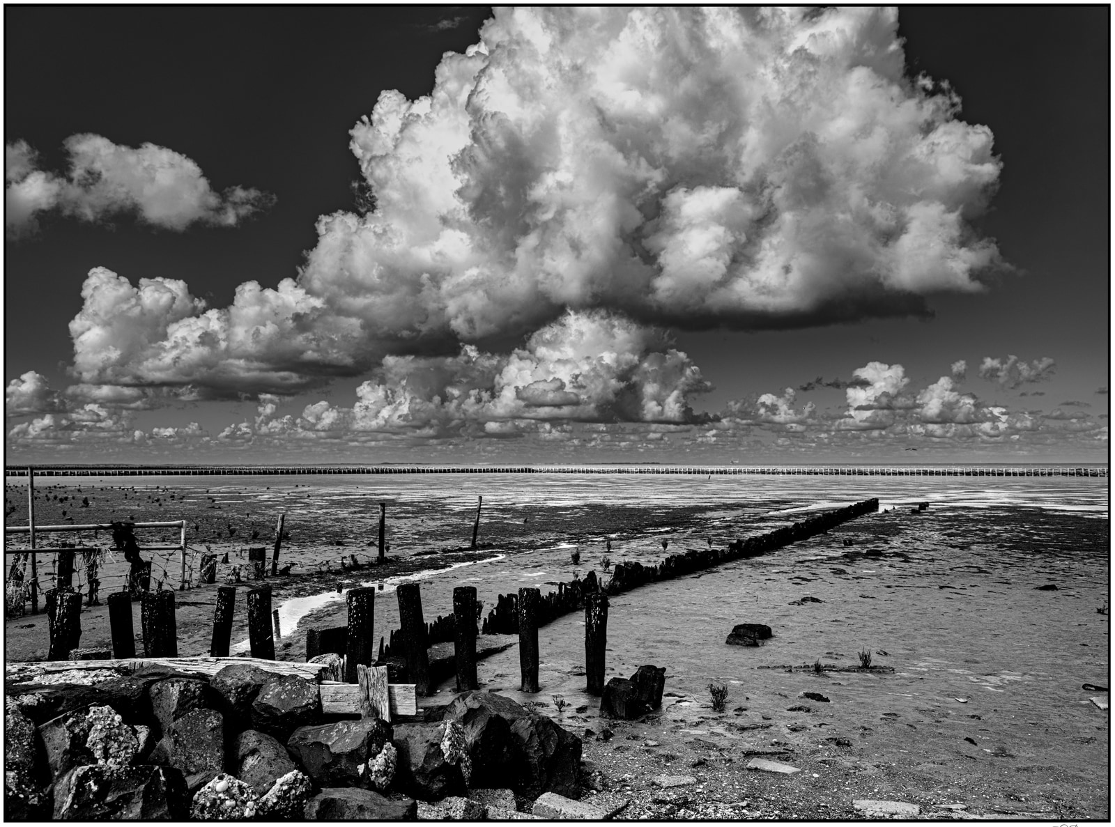 A wide, flat landscape stretches under a dramatic sky filled with towering cumulus clouds; wooden posts and scattered rocks line the foreground.