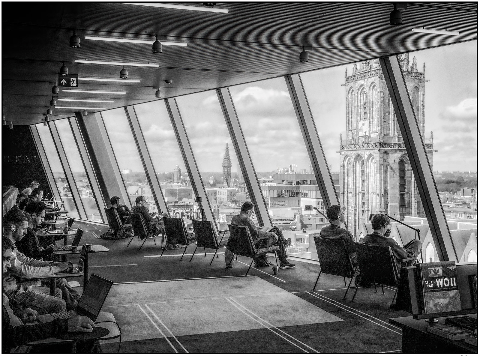 People sit at tables with laptops in a modern, high-rise lounge, looking out through large windows at a cityscape with a prominent Gothic tower.