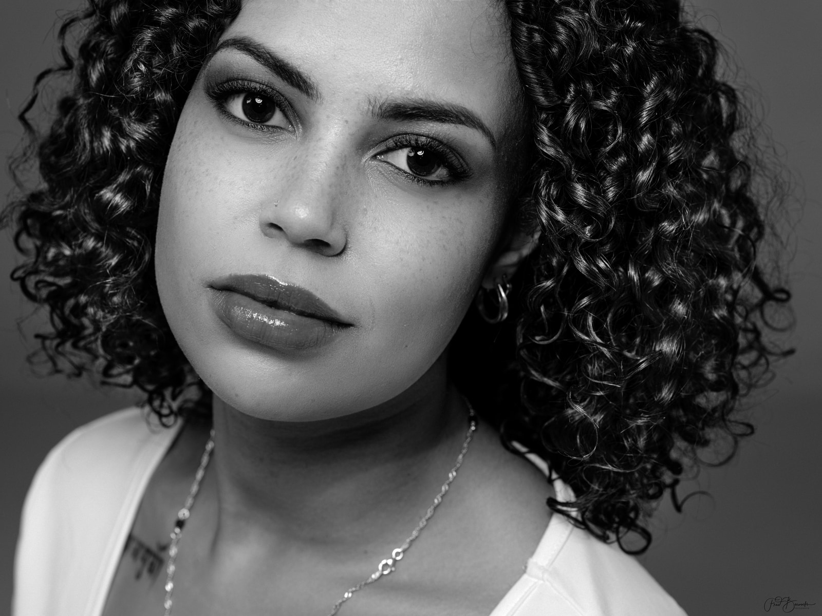A close-up portrait of a person with curly hair, wearing a light-colored top and a delicate necklace, set against a dark background.