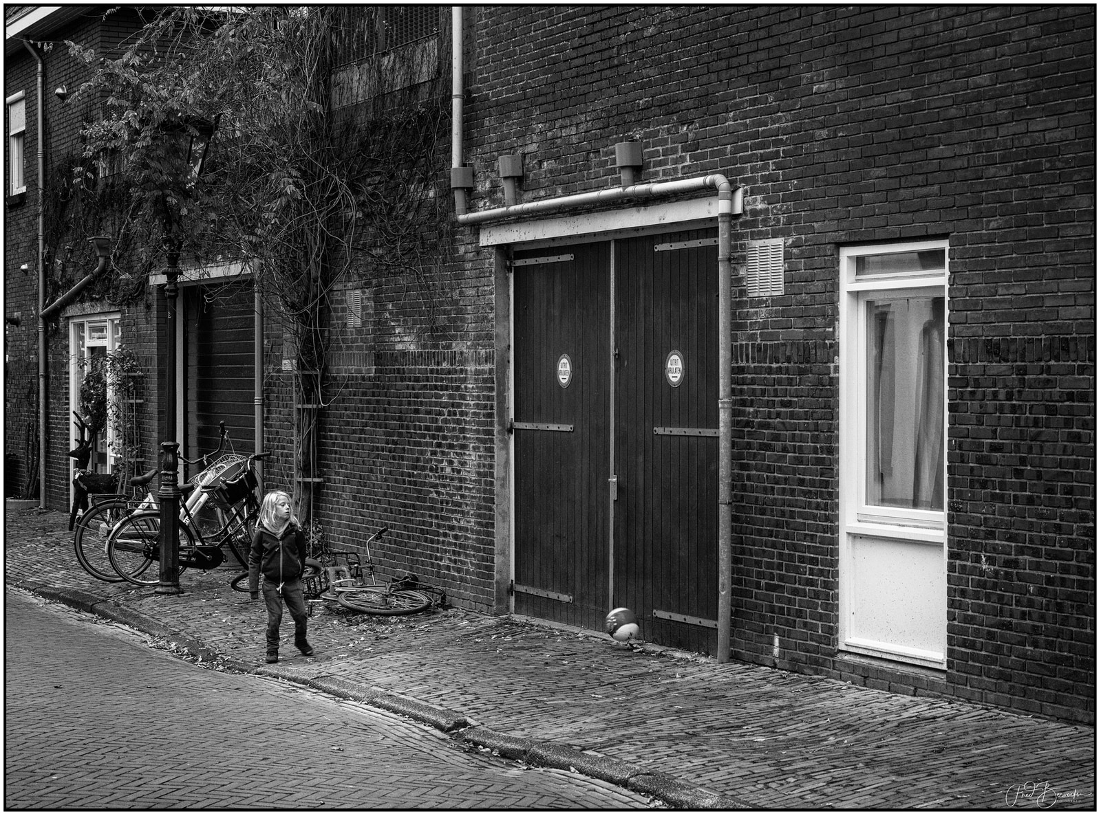 A child stands near bicycles beside a brick building with a large wooden door and a window.