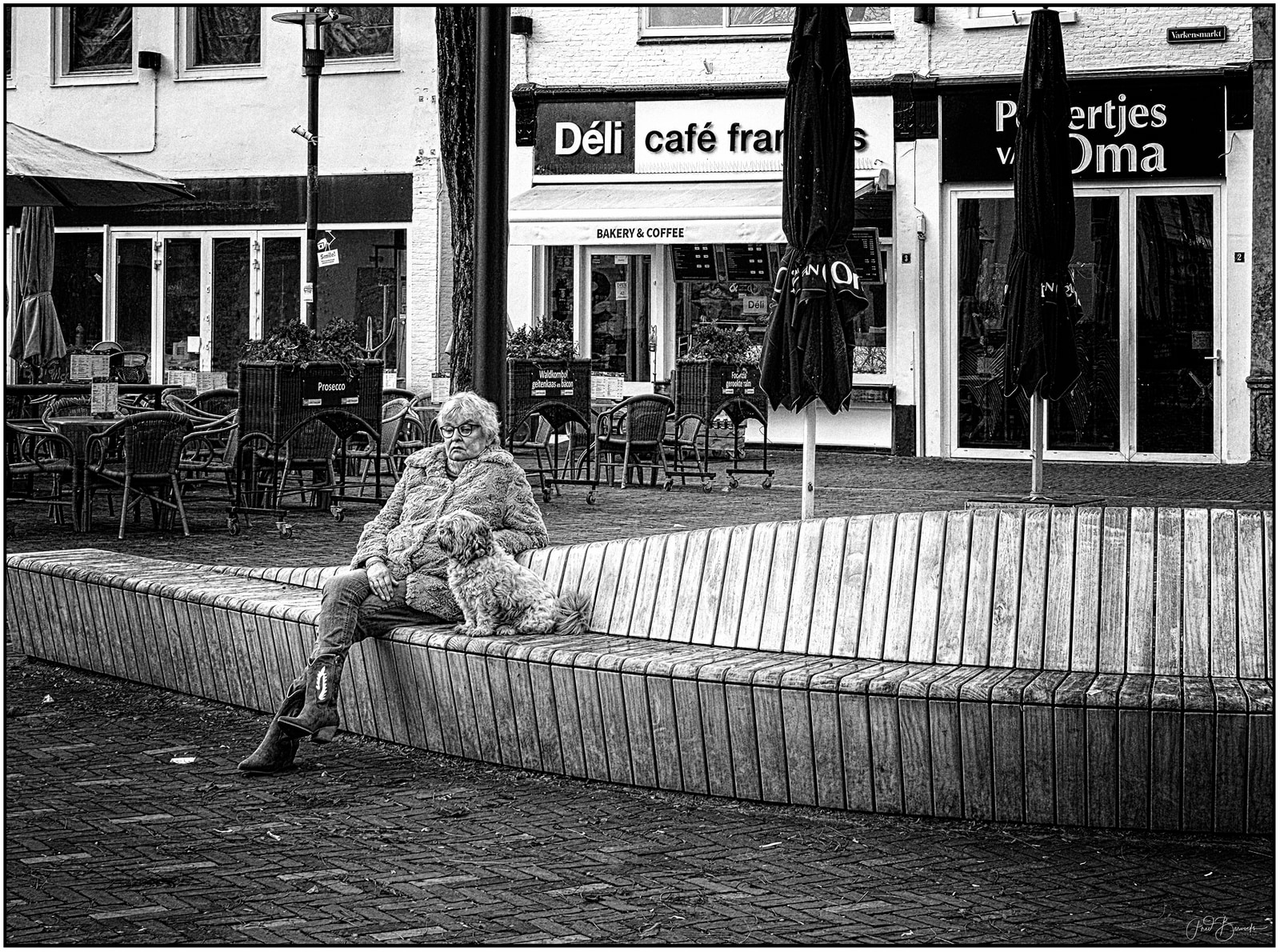 An older woman sits on a long bench with a small dog beside her, in front of a café with outdoor tables and closed umbrellas.